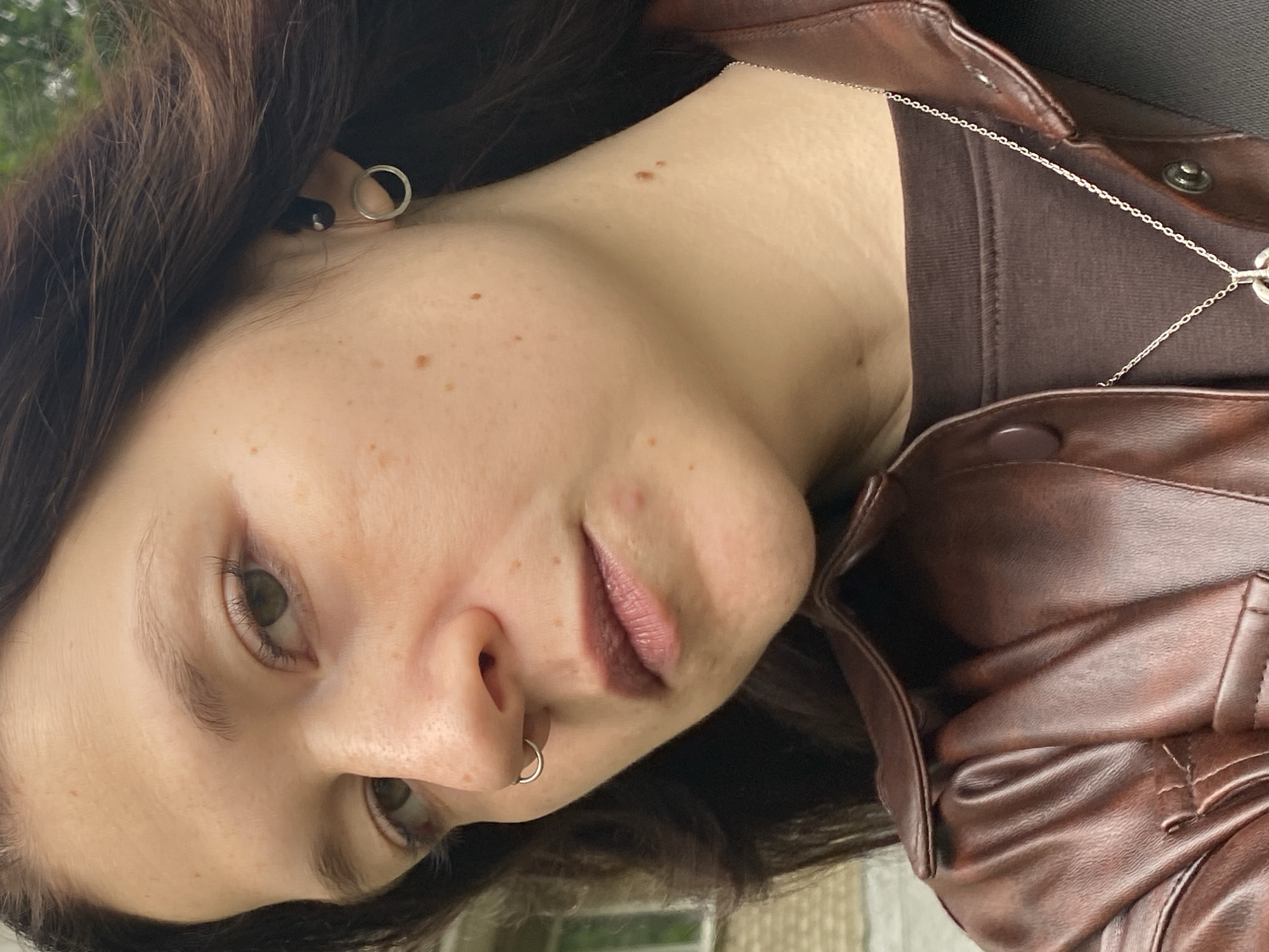 A close-up, angled portrait features a young person, likely female, with dark brown hair and light skin, positioned in what appears to be an outdoor setting in Chaikovsky, Russia. The subject is looking directly towards the camera with a neutral expression. They have greenish-brown eyes and a smattering of freckles visible across their face, neck, and chest.
They are wearing a dark brown, possibly long-sleeved, top underneath a brown leather or faux-leather jacket. Accessories include a small silver hoop earring in their right ear and a black stud in their left. A silver septum ring is visible in their nose. A thin silver chain necklace hangs around their neck, with a small, indistinct pendant resting on their chest, partly obscured by the jacket's collar. The jacket has visible buttons or snaps.
The background is softly blurred, showing hints of green foliage and a light-colored structure, possibly a brick or concrete building, suggesting a daytime scene outdoors. There is no visible text in the image.