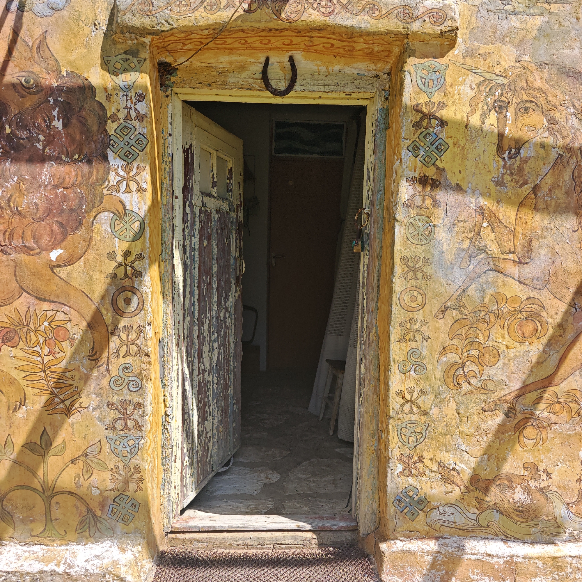 An open, severely weathered wooden doorway pierces a stucco facade, richly decorated with faded and peeling murals, located in Останинское сельское поселение, Ukraine. The facade exhibits a predominantly yellow and ochre palette with underlying layers of brown and white paint.
On the left side of the doorway, a large, stylized brown creature, resembling a lion or bull with a prominent eye and shaggy mane, is painted. Below it, branches with pomegranates and foliage are depicted. A vertical column of patterned symbols, including circular motifs, stylized plant forms, a triskelion, and interwoven knot designs, runs along the left door jamb.
The right side of the doorway features a unicorn-like creature with a horn, flowing mane, and a human-like eye. Below this, a large, fish-like or dragon-like creature emerges from waves, exhaling a smaller fish. Further plant motifs are present. A corresponding vertical column of symbolic patterns, mirroring those on the left, adorns the right door jamb.
The wooden door frame is heavily distressed, with multiple layers of paint (yellow, brown) flaking off. A rusty, inverted horseshoe is nailed centrally to the wooden lintel above the doorway, which also bears faded geometric patterns. The wooden door itself, painted white with red and brown undertones, is ajar, revealing significant paint loss and exposed wood grain. A dark, woven mesh mat lies on the ground directly in front of the worn wooden threshold.
Through the open doorway, a dimly lit interior is visible. Another plain, dark brown door is situated further within the space. To the right of the interior view, a roll of white mesh material leans against a wall next to a small wooden stool. The interior floor is uneven and appears to be earth or worn concrete. A dark, rectangular object, possibly a framed picture or a covered window, is mounted above the inner doorway. Strong sunlight illuminates the exterior facade, casting sharp shadows from the door frame onto the painted walls, highlighting the texture of the deteriorating surfaces.