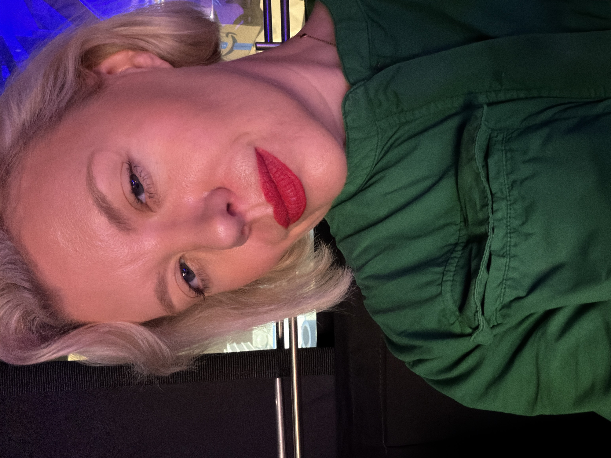 The image features a close-up, sideways portrait of a blonde woman with fair skin and red lipstick. She is wearing a dark green garment, possibly a jacket or shirt, with visible stitching and a pocket detail. Her hair is styled in soft waves. The lighting in the image appears to be artificial, casting a warm, reddish-pink hue on her face. The background is dark and out of focus, with some vertical elements that could be furniture or a room divider. There are no visible signs or text. The overall impression is a casual, self-taken photograph, likely indoors, given the lighting and background. The woman's expression is neutral. The context provided indicates the location as Culver City, USA.