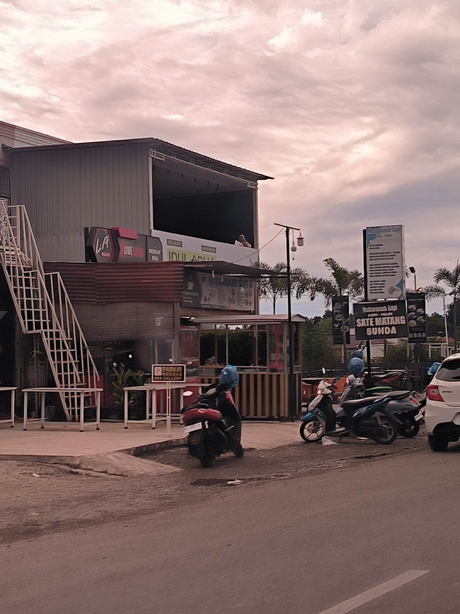The image presents an outdoor street scene in Keuniree, Indonesia, under an overcast sky, likely in the late afternoon or early evening due to the soft, warm light. The central feature is a two-story commercial building with an industrial aesthetic, characterized by corrugated metal walls. An external white metal staircase with railings provides access to the upper level, where a person is subtly visible on an open-air balcony.
On the ground floor, an outdoor seating area with white tables and chairs is set up. Several motorcycles of various colors, including red, blue, and black, are parked on the paved area in front of the building. A white car is partially visible on the far right. Palm trees dot the background, hinting at a tropical climate.
Various signs advertise businesses. Prominently displayed text includes "LA," "LIVE," and "BERKAT DUA" on signs attached to the building. Another sign directs attention to "SATE MATANG BUNDA," likely a food establishment, with "MALEM" partially visible. A smaller sign clearly indicates "PARKIR KHUSUS MMA GALLERY" (Special Parking MMA Gallery). Other visible text snippets include "SELECT" and "IDUL ADHA." The scene conveys a quiet, daily street ambiance with commercial activity.
