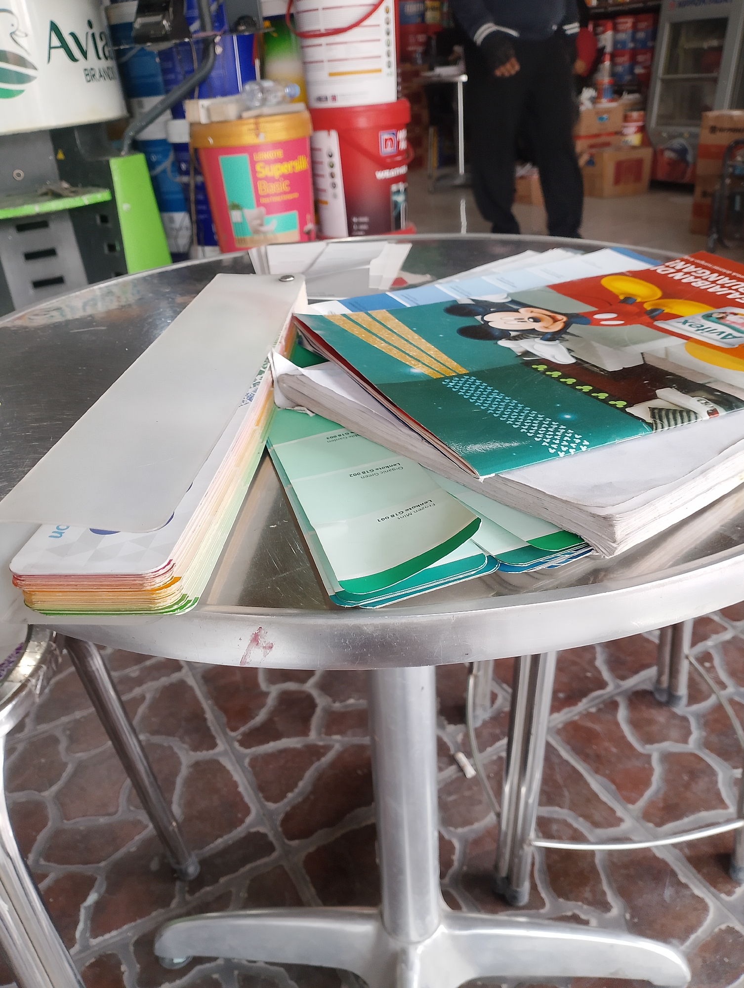 An overhead shot captures a stack of paint swatch books on a metallic, circular table, situated in an indoor retail environment. The books, fanned out, reveal colorful hues and text, with one prominently featuring Mickey Mouse graphics. The table itself is supported by slender metal legs, contrasting with the reddish-brown tiled flooring. In the background, shelves are laden with various paint cans, and a blurry figure, dressed in black, stands behind the table. A partially visible white plastic container with a pink label that reads "SuperSilk Basic" is positioned to the left of the table. The scene is likely a paint store or hardware shop in Blimbing, Indonesia, as indicated by the store's products.