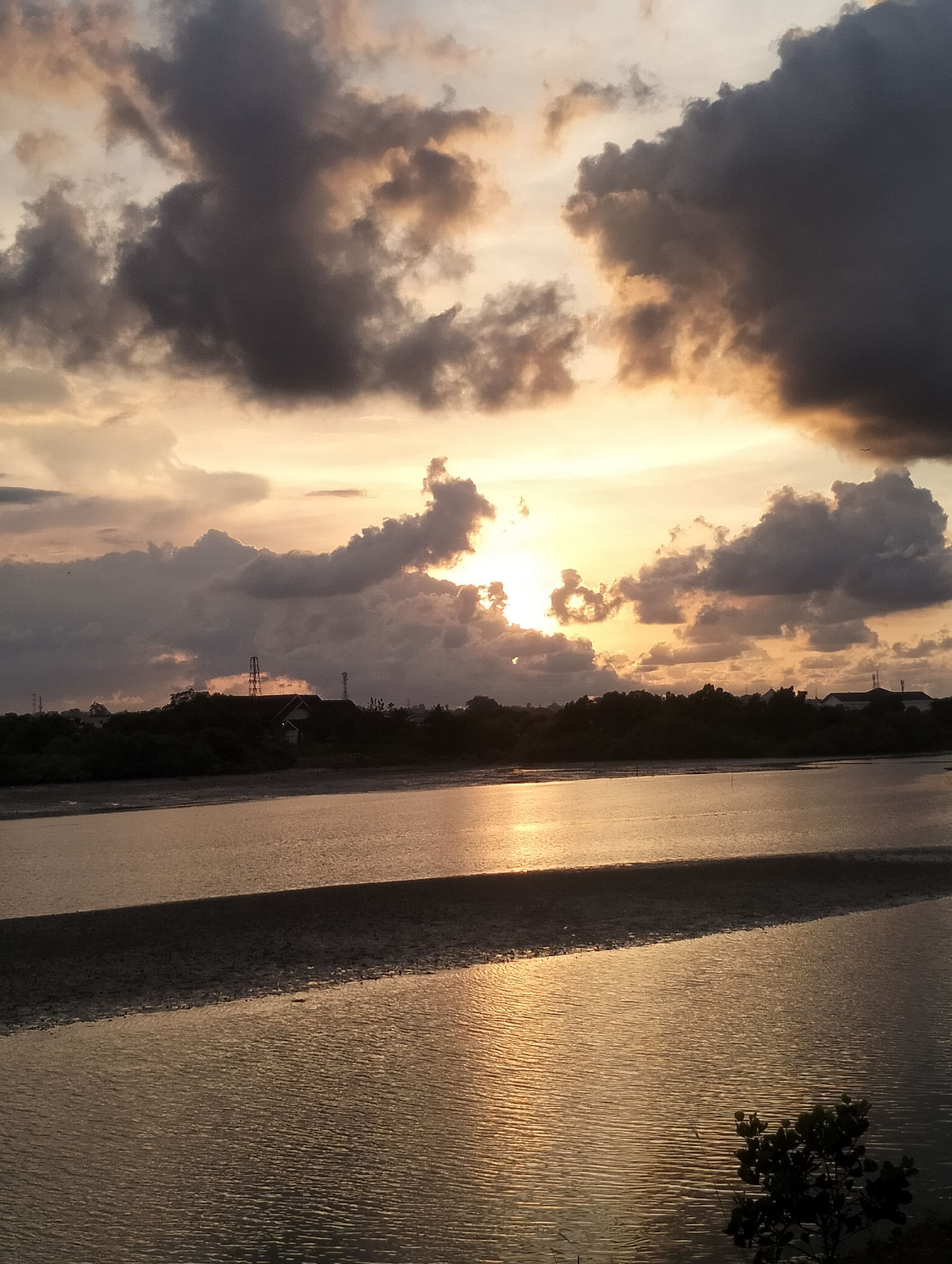The image depicts a sunset over a body of water, possibly a river or bay. The sky is filled with dark, dramatic clouds, with the sun visible through a break in the clouds. The sunlight reflects on the water's surface, creating a golden glow.
In the background, there is a strip of land with trees and some buildings or structures, potentially part of the city of Lhokseumawe in Indonesia. The foreground shows the water with some texture or ripples on the surface, and a small tree or bush visible in the lower right corner. There is no explicit information or detail within the image to confirm specific location in the city, but could possibly be near a coastal area.
The scene is devoid of people. The primary subjects are the sky, clouds, sun, water, and the distant land. The action is the natural occurrence of a sunset. There is no evidence of interaction, this appears to be a captured moment in nature.