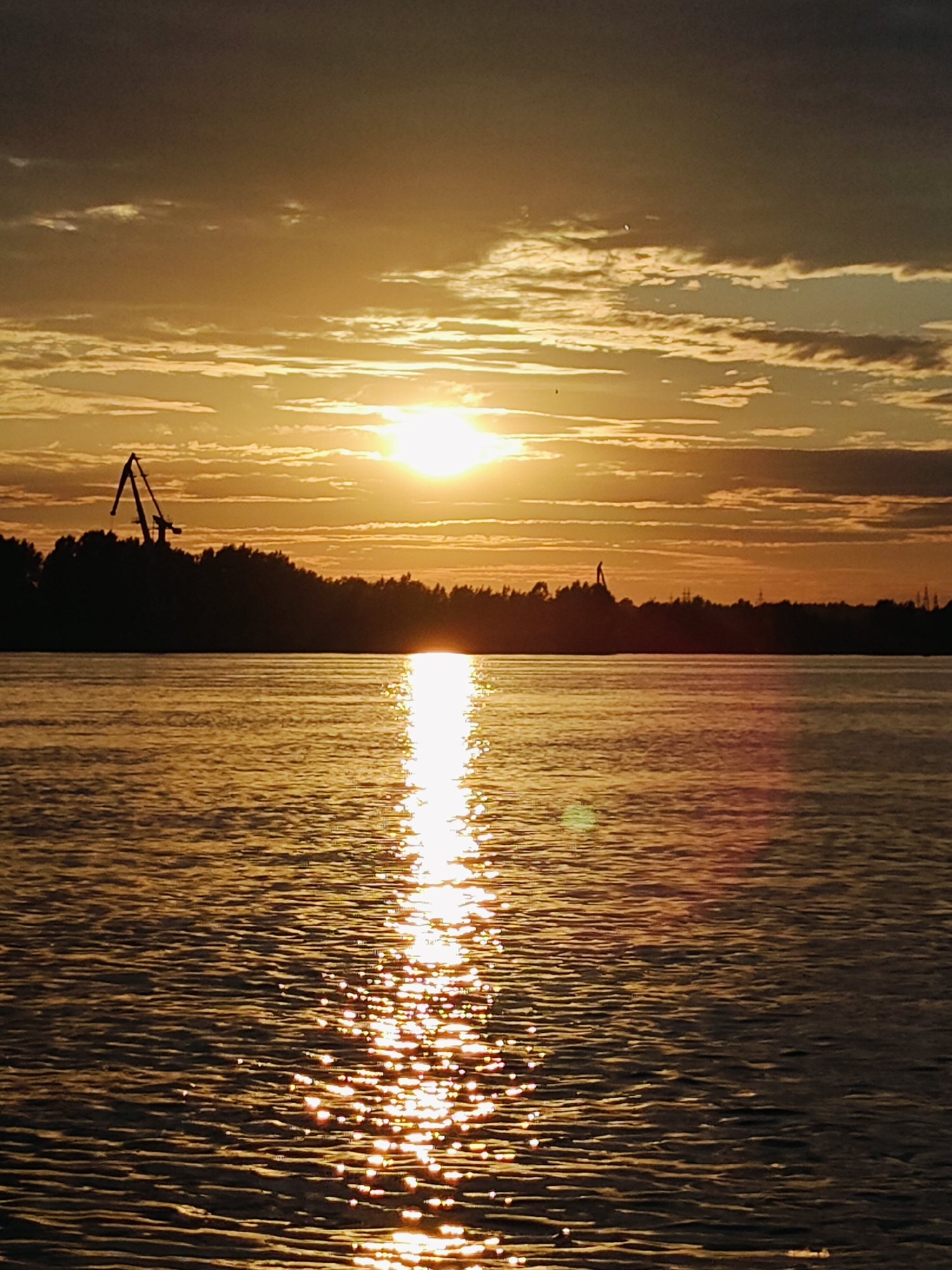 This image captures a serene sunset over a body of water, likely a river, in Chaikovsky, Russia. The sky is filled with dramatic clouds, tinged with warm hues of orange and yellow as the sun descends. A bright, radiant sun is visible near the center of the horizon, casting a shimmering reflection on the water's surface. The water itself appears calm, with gentle ripples reflecting the golden light of the sunset.
Silhouetted against the vibrant sky is a dense treeline, marking the distant shore. On the left side of the image, a large industrial crane stands out in silhouette, suggesting a port or industrial area adjacent to the natural landscape. Further in the distance, another, smaller crane or structure is faintly visible. The overall atmosphere is one of peaceful tranquility, with the natural beauty of the sunset contrasting with the industrial elements on the shore. The time of day is clearly evening, as indicated by the sunset. The weather appears clear with scattered clouds. No text is visible in the image.