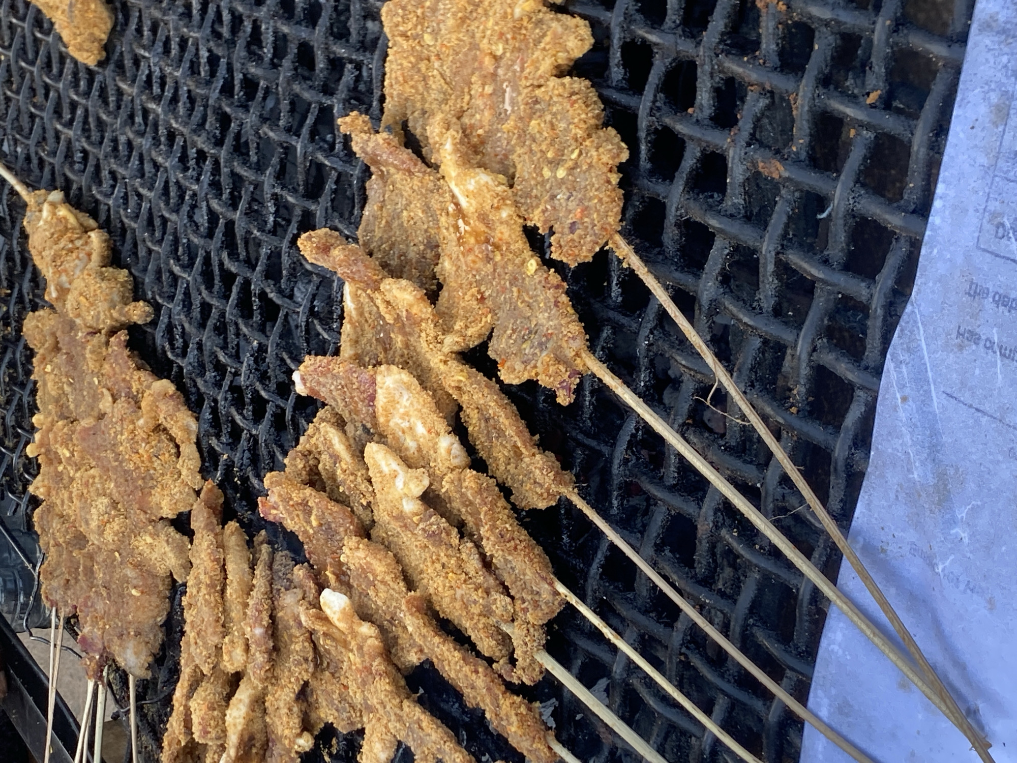 The image shows skewers of meat, likely beef, coated in a grainy, reddish-brown spice blend, resting on a black wire grill. The skewers are arranged in a cluster, with some extending off the grill. A piece of white paper is visible to the right of the grill. Given the prompt's location in Bukuru, Nigeria, it is likely street food being prepared for sale.