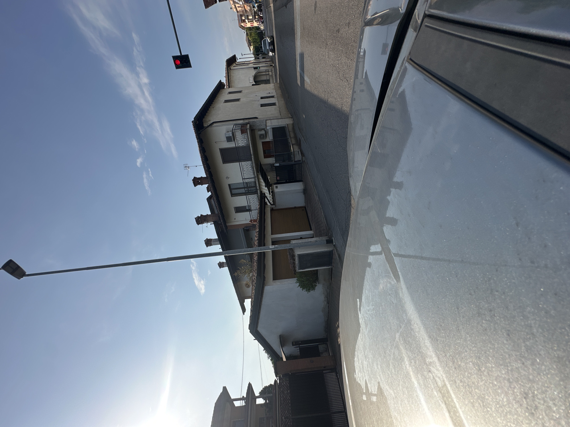 The image depicts a street scene in Rudiano, Italy. The perspective is from inside a vehicle, looking forward and slightly upwards. The foreground shows the hood of a light-colored car, dusted with a fine layer of particles, reflecting the sky and surrounding buildings.
In the mid-ground, a paved road curves to the right, with houses and a garage visible on the left and right sides. The houses appear to be two-story residential buildings with tiled roofs and balconies. One house on the left has a dark metal gate and a brick pillar. Another house to the right, painted white, features multiple windows and balconies. Adjacent to this house is a garage with a light brown, segmented door. Further down the street, other buildings and trees can be seen.
In the background, the sky is a clear blue with some wispy white clouds and a bright sun is visible on the left side. A tall, dark lamppost stands prominently on the left side of the street. A traffic light with a red illuminated signal is positioned above the road on the right side, attached to a horizontal arm extending from a pole. A dark-colored car is driving on the road in the distance, approaching the intersection controlled by the traffic light. The overall impression is a sunny day in a residential area.