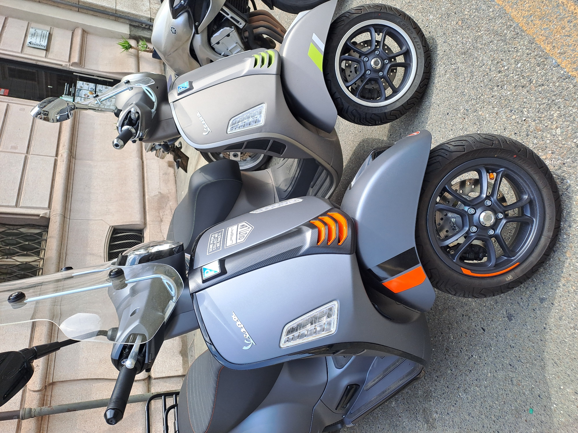 The image shows two parked scooters in front of a building in Turin, Italy. The scooter in the foreground is a gray Vespa with orange accents on the front fender and wheel rim. It has a transparent windshield and black handlebars. A sticker on the front of the scooter reads "SOME PEOPLE COLLECT ART - WE RACE IT - URBAN OUTLAW". The scooter behind it is also a gray Vespa, with a silver Yamaha motorcycle visible to the right. The building has a stone facade with arched windows and a closed doorway. The ground is paved with asphalt.