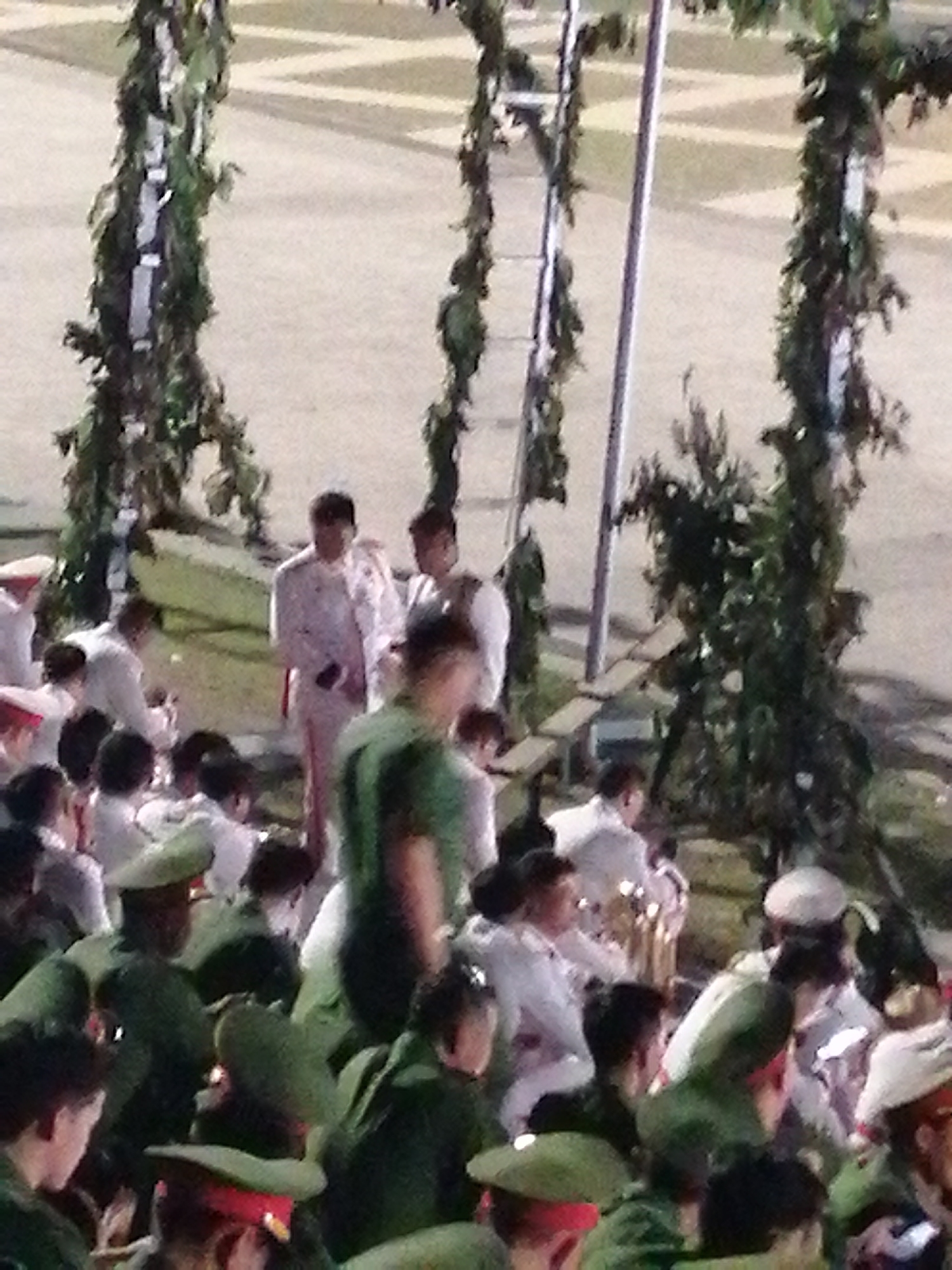 The image shows a gathering of people, many of whom appear to be in uniform, with some wearing white formal attire and others in green military-style uniforms and hats. The scene is set outdoors, with decorative green foliage structures that resemble arches or columns framing the left and right sides of the image. In the background, there are hints of a paved area with geometric patterns. The lighting suggests it might be late afternoon or early evening. The overall impression is of a formal event or ceremony. Based on the context of the provided crops, this scene is likely taking place in Da Lat, Vietnam, a city known for its French colonial architecture and lush landscapes, where such events might occur.