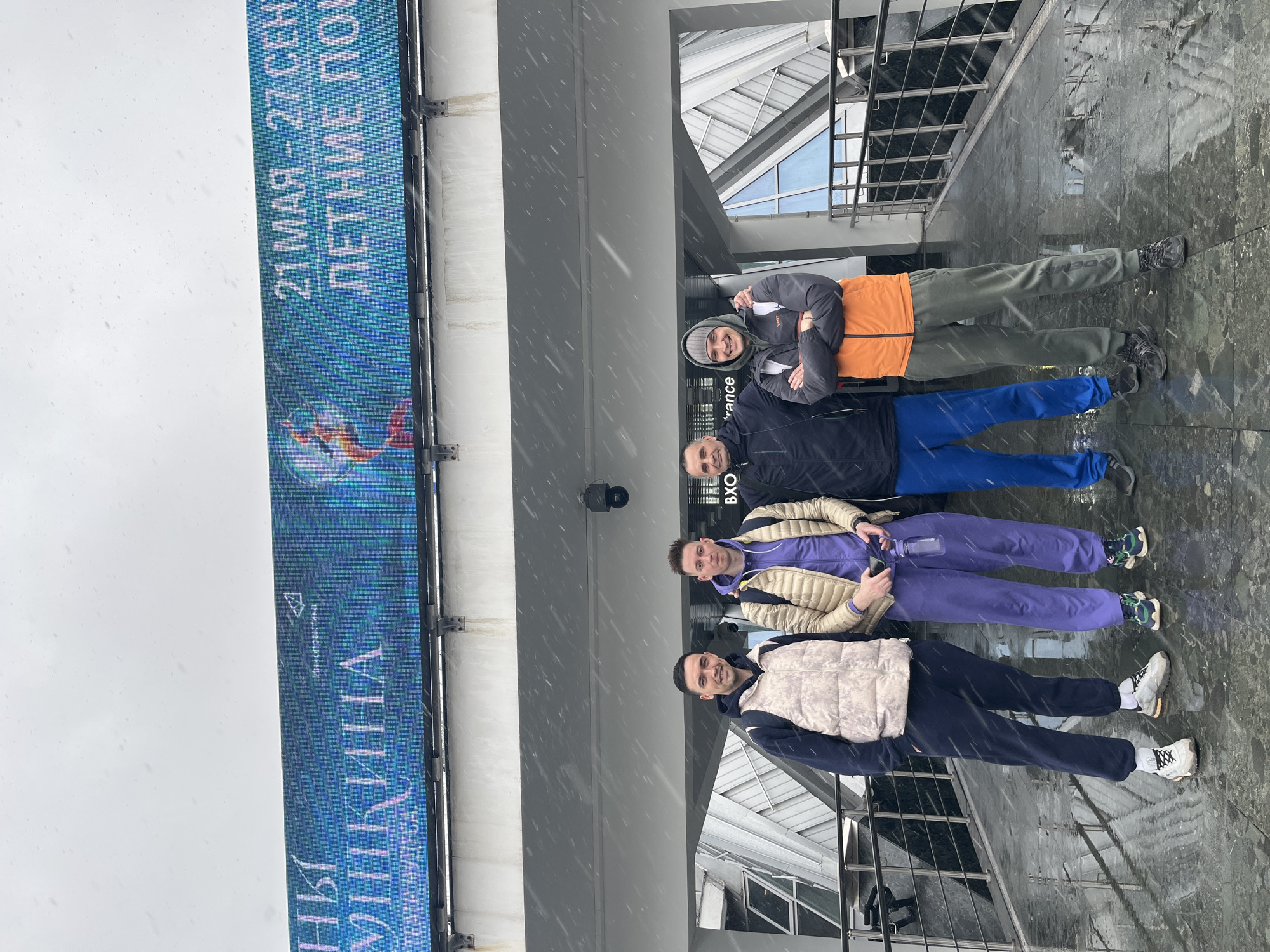 Here is a description of the image:
Four men are standing in a row, posing for a photo near a large digital display in Moscow, Russia. The display advertises "NI UNIKINA" at the "TEATR CHUDESA" (Theater of Miracles), with dates "21 MAY - 27 SEPT" and the phrase "SUMMER". The men are dressed in casual clothing: the first man is wearing a white vest, the second is wearing a tan jacket, the third is wearing a blue jacket, and the fourth is wearing an orange jacket. The ground is wet, indicating rain or snow. The backdrop includes architectural elements of a modern building with glass windows and a metal structure. It appears to be snowing.