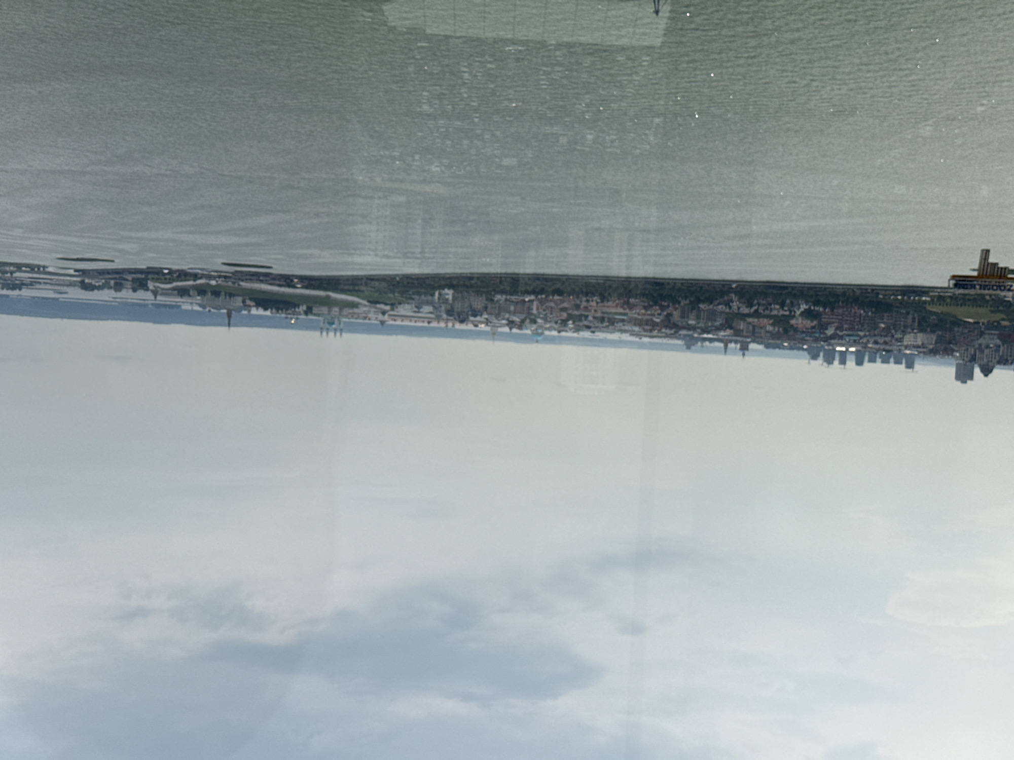 The image shows an aerial view of the city of Kazan, Russia, looking across a body of water towards the city skyline. In the foreground is a large expanse of green-tinged water with subtle ripples on its surface. In the mid-ground, a long, densely populated cityscape stretches along the waterfront, characterized by a mix of architectural styles. Prominent features include numerous buildings, some with domed roofs, and a line of trees bordering the water. On the left side of the image, a section of a pier or dock structure with the word "ZOOMER" is visible. In the background, the sky is overcast with soft, muted clouds, suggesting a hazy or slightly overcast day. The overall impression is one of a panoramic urban landscape viewed from a distance across a wide waterway.