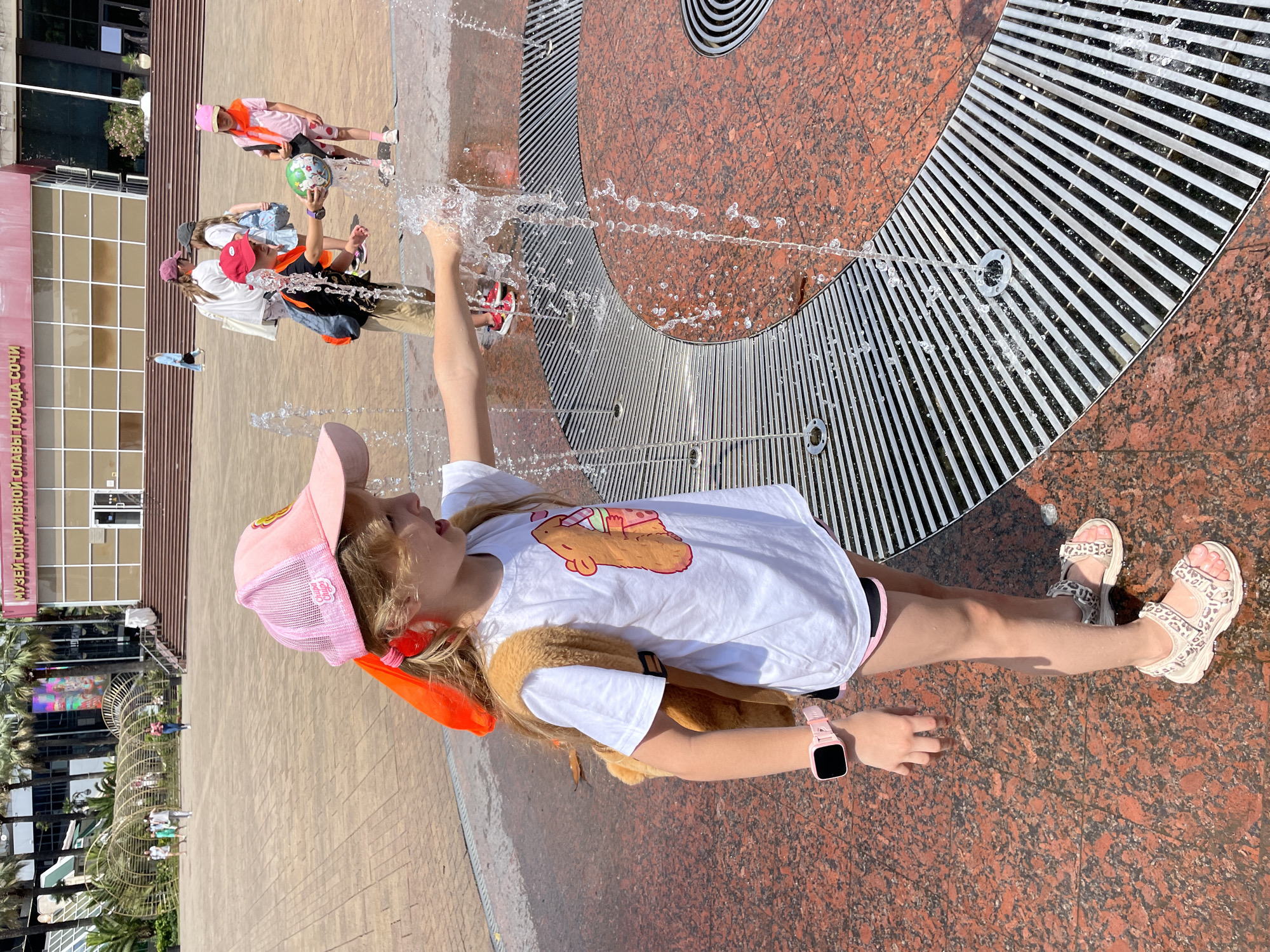 In a sun-drenched outdoor setting, a young girl is captured mid-action, playing in a fountain. She is wearing a pink baseball cap, a white t-shirt with a cartoonish brown animal graphic, and animal-print sandals. A bright orange scarf is tied around her ponytail, and a pink smartwatch adorns her wrist. Her legs are bent, and water sprays around her feet, suggesting she is actively engaged with the water jets.
In the background, several other children are visible, also interacting with the fountain. One child, further back, is wearing a pink outfit and holding a colorful ball. The environment appears to be a public plaza with a patterned granite floor and metal grates characteristic of a water feature. The bright sunlight indicates it is daytime, and the overall mood is one of playful enjoyment.
To the left of the frame, a building with a modern facade and glass windows is partially visible. A sign on the building, written in Cyrillic script, reads "МУЗЕЙ СПОРТИВНОЙ СЛА..." which likely translates to "Museum of Sport...". Palm trees are also visible in the background, hinting at a warm climate. The scene takes place in Sochi, Russia, as indicated by the location context.