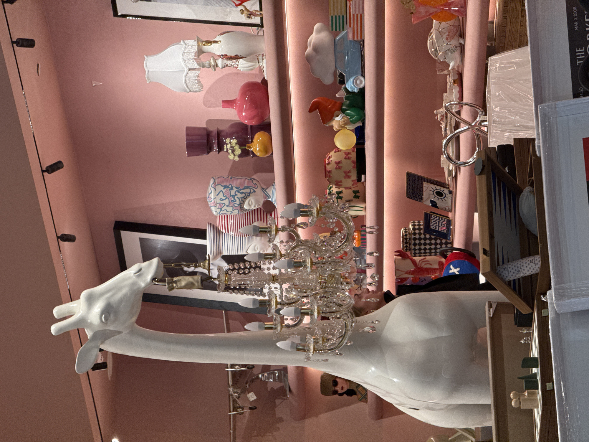 The image depicts a retail space in Belgrade, Serbia. A white, ceramic giraffe statue with a chandelier hanging from its neck serves as a prominent feature. The background is a pink wall displaying shelves filled with various decorative items, including vases, figurines, and what appears to be a game board. A person with sunglasses and a plaid shirt is partially visible in the lower center, appearing to be looking at the merchandise. The store has an eclectic and artistic vibe.