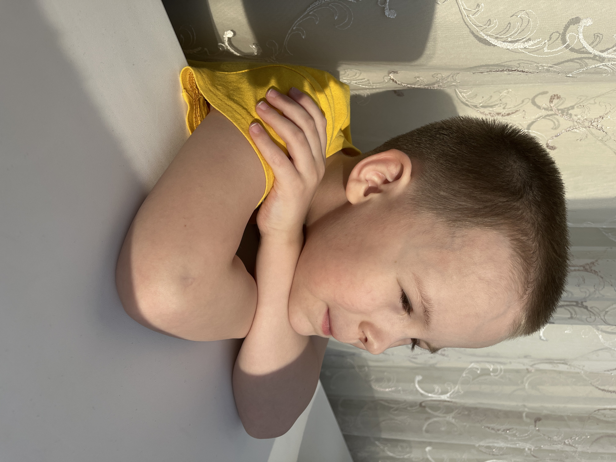A young boy with short, dark hair is resting his head on his arms, which are crossed on a white table. He is wearing a yellow shirt. The background features a patterned white curtain. The location is Usman, Russia.