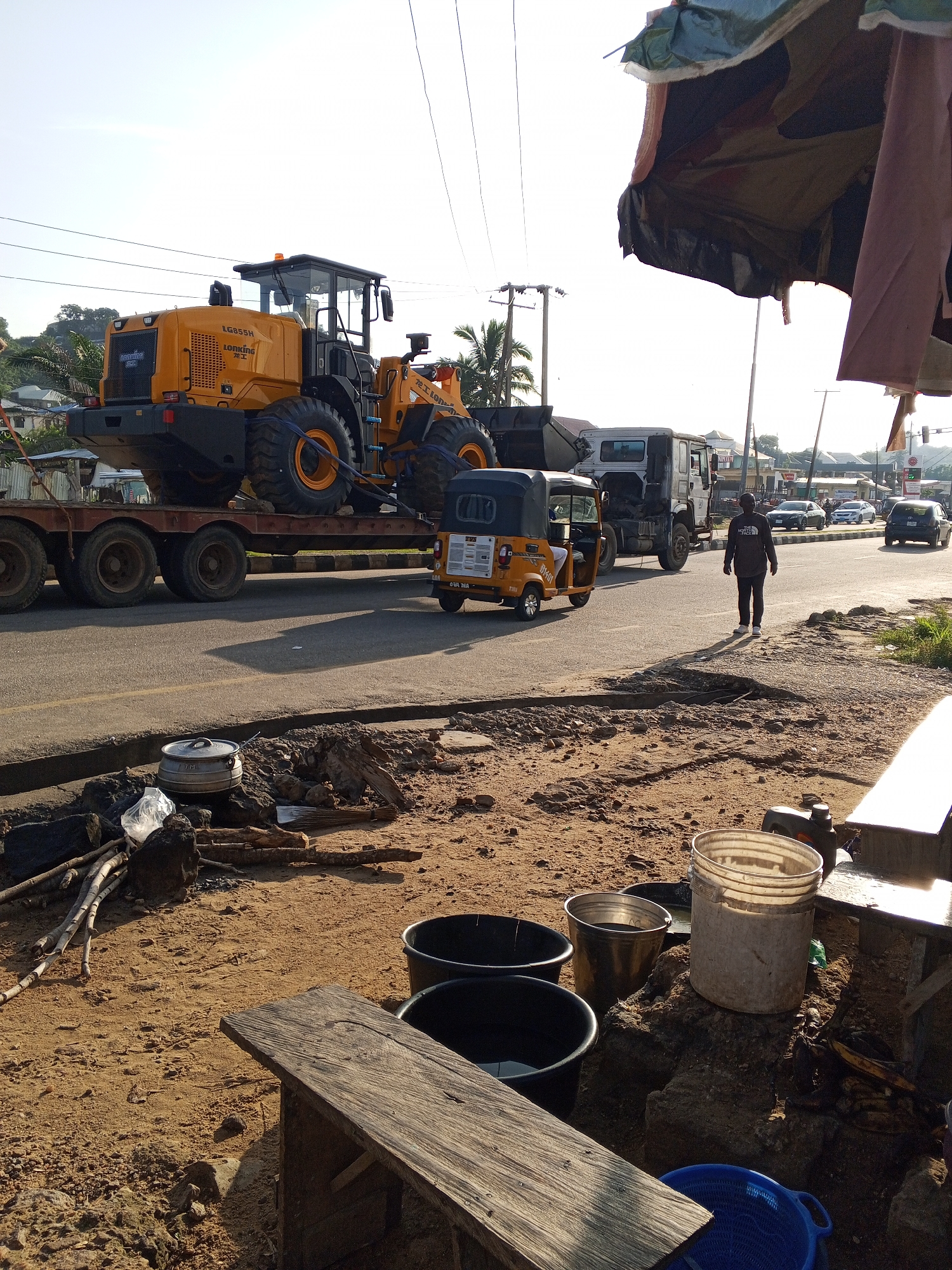 The image depicts a street scene in Lokoja, Nigeria. The primary subjects are a large yellow front-end loader, being transported on a flatbed trailer, a white truck, and a yellow auto-rickshaw. A man is walking away from the camera on the right side of the road. Various objects are scattered on the dusty ground in the foreground, including buckets, logs, rocks, and a pot.
The front-end loader is a prominent feature, with the brand name "LG" and model "LG855H" visible on its side, along with "LONKING" written below it. The auto-rickshaw has text on its side, including what appears to be "OKP" and "NMA". The man walking on the right is wearing a dark jacket with a logo on the back that reads "THE NORTH FACE".
The scene appears to be set during the daytime, with clear skies and long shadows indicating either early morning or late afternoon. The lighting suggests a sunny day. The presence of the construction equipment and the overall dusty, unpaved area along the roadside suggest a possibly developing or uncompleted infrastructure area. The arrangement of buckets and other items on the ground hints at informal commerce or daily life activities occurring roadside. The overall atmosphere is that of a typical busy street in a Nigerian city, with elements of both commercial transport and everyday life.