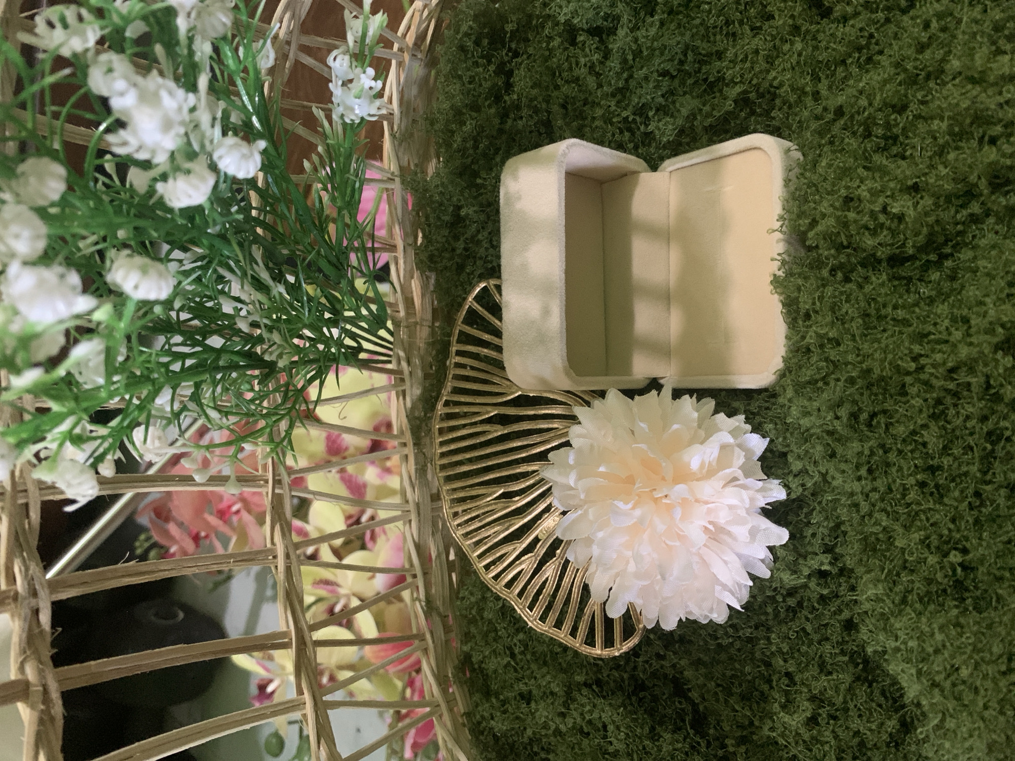 In Kuala Pembuang, Indonesia, a close-up shot captures a decorative arrangement on a bed of artificial green moss. The composition features an open, beige velvet ring box, its interior lined with cream-colored fabric, positioned slightly to the right of center. To the left of the ring box rests a delicate, pale pink artificial flower with layered petals, positioned in front of a gold, fan-shaped decorative element with radiating lines. In the background, a woven basket made of light-colored reeds contains a cluster of white artificial flowers with green stems and foliage, as well as some blurred pink and yellow flowers. The overall scene is static and appears to be set up for a display or event.