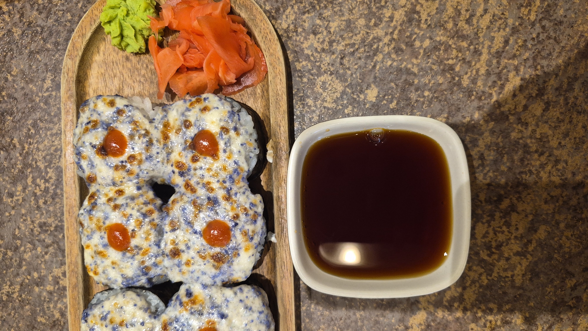 The image shows a close-up, overhead view of a sushi platter. The sushi rolls are topped with a white sauce that has been broiled, with some blue specks and golden-brown crispy bits. Each piece of sushi is garnished with a small dollop of bright red sauce. To the right of the sushi, there is a pile of thinly sliced pink pickled ginger and a small mound of bright green wasabi. The sushi and accompaniments are arranged on a light-colored wooden platter. In the foreground, there is a white square dish filled with dark brown soy sauce. The entire scene is set against a speckled, dark brown countertop. There are no people visible in the image. The location is not specified in the image.