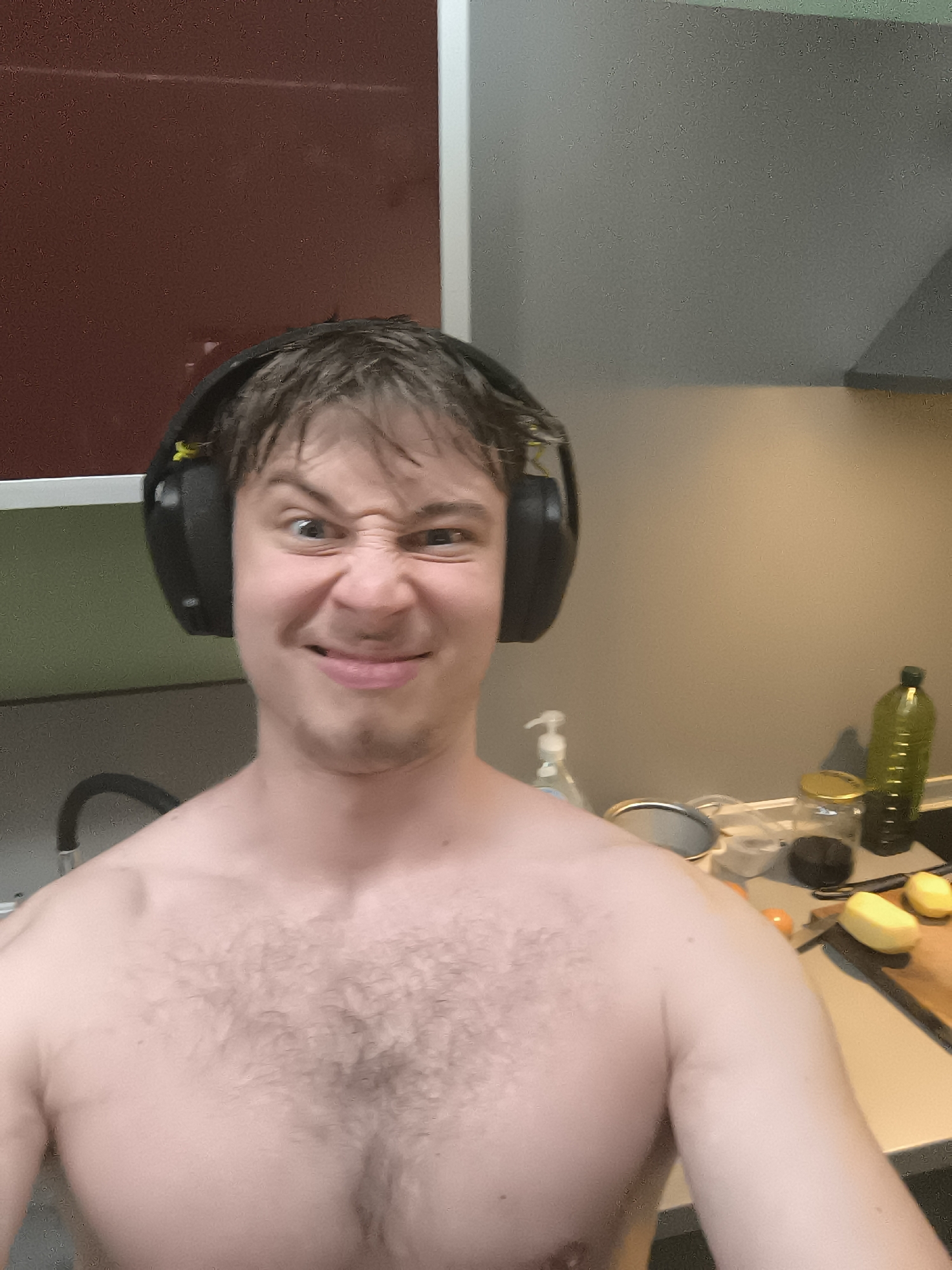 A shirtless man with short, wet hair and black headphones is pulling a humorous face at the camera. He is standing in a kitchen with red and white cabinets in the background. On the counter to his right, there is a sink faucet, hand soap, a round mirror, a jar with a gold lid, a bottle of olive oil, a cutting board, a knife, and some potatoes. The lighting is somewhat dim. The location is not discernible as Sofia, Bulgaria from the visual information provided.