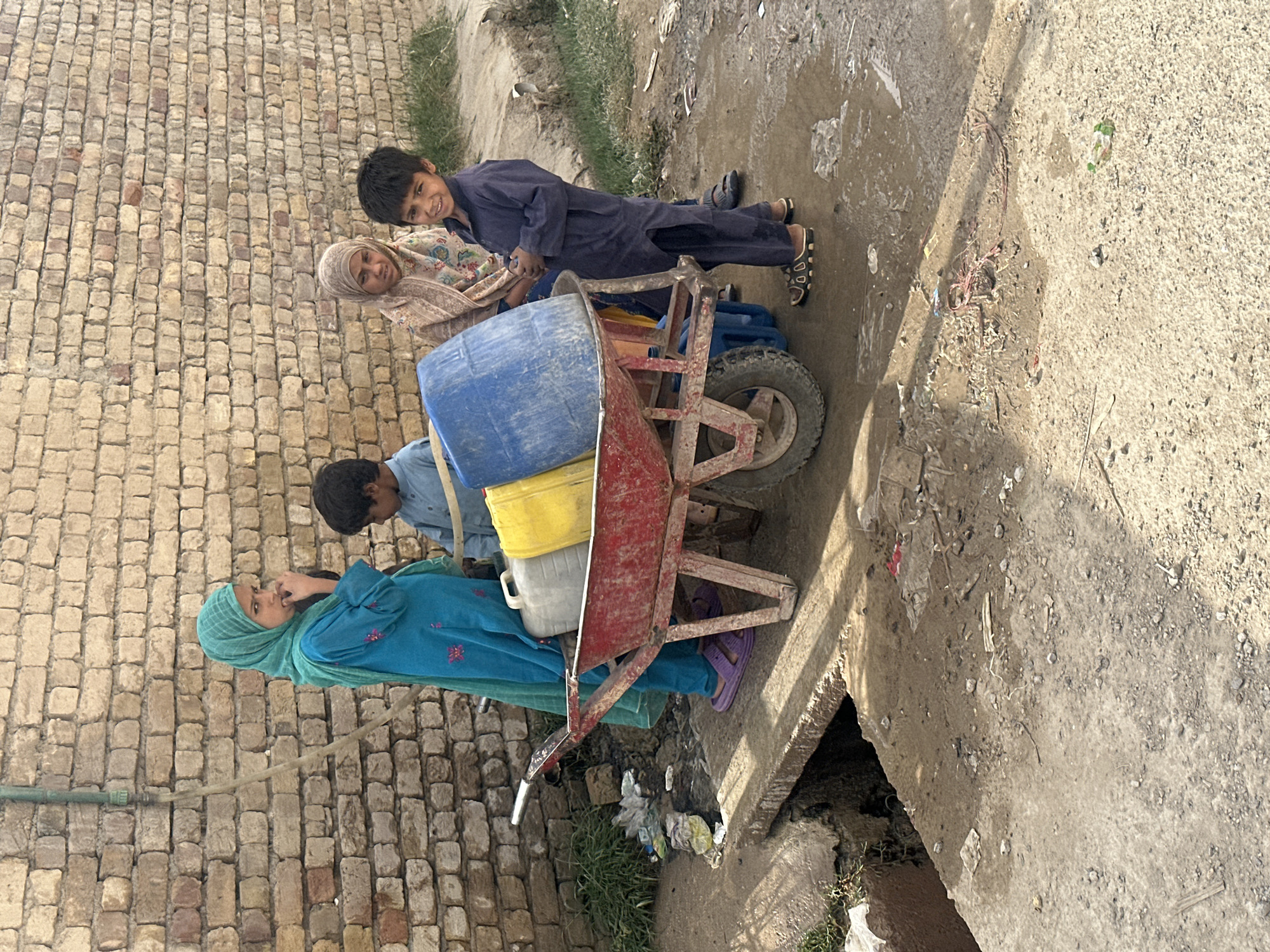 In Quetta City Tehsil, Pakistan, a woman wearing a blue headscarf and blue clothing is pushing a red wheelbarrow loaded with a blue barrel, a yellow container, and a white container. A boy in a blue outfit stands beside the wheelbarrow, looking at the camera. An older woman wearing a patterned headscarf and shawl stands behind the boy, also looking toward the camera. The ground is partially paved and has patches of dirt and scattered debris. A brick wall is visible in the background.