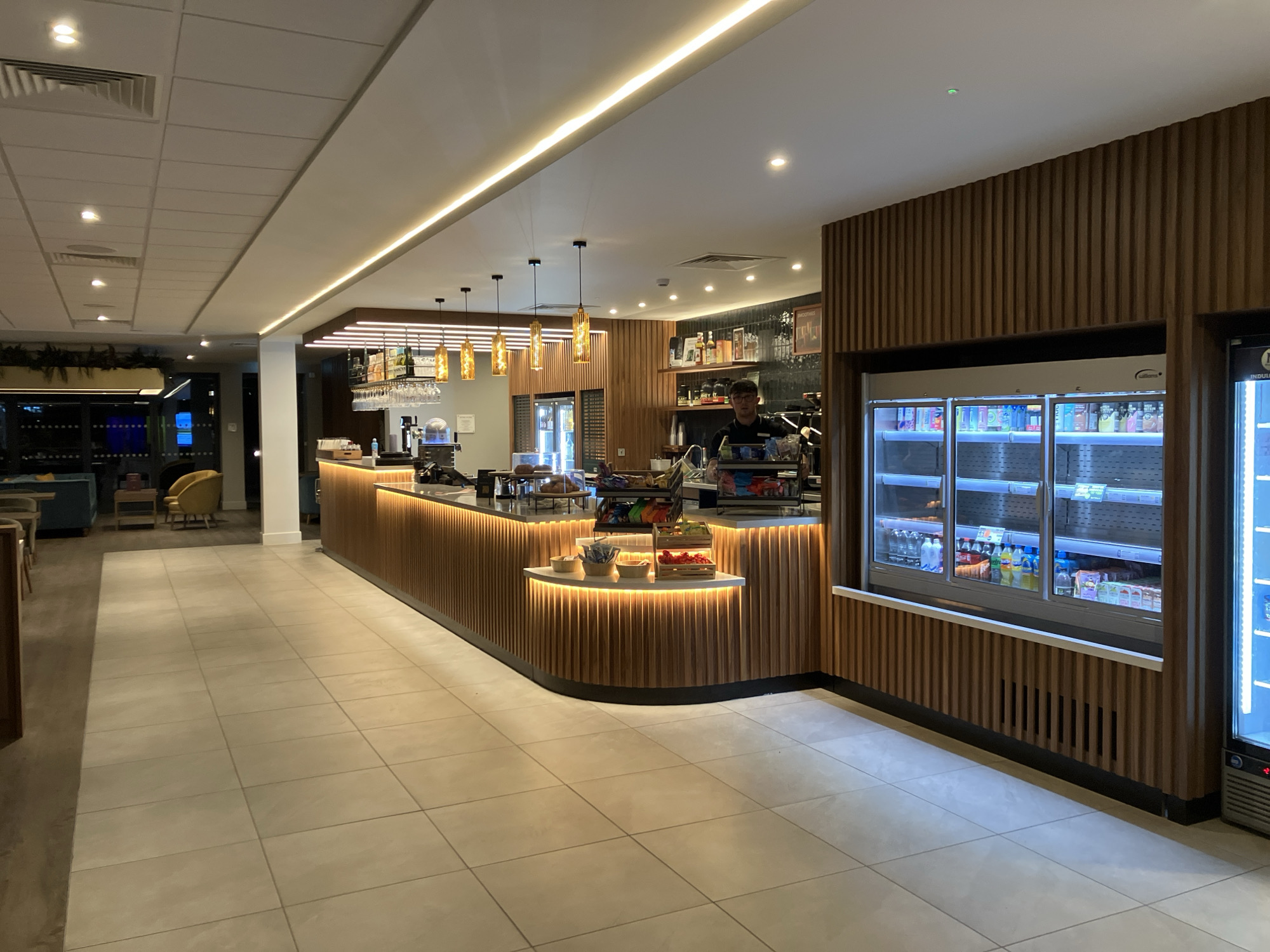The image depicts a well-lit indoor space in West Suffolk, United Kingdom, likely a café or lounge area. The scene is focused on a curved counter with a wooden panel design and integrated lighting. Behind the counter, a person is visible, possibly a barista, standing amidst coffee-making equipment and shelves stocked with items. Displayed on the counter are assorted pastries, snacks, and fresh fruit. To the right of the counter, built-in refrigerated display cases showcase beverages. The floor is covered in square tiles. The ceiling has integrated lighting and a modern design. In the background, seating areas with blue and yellow furniture can be seen.