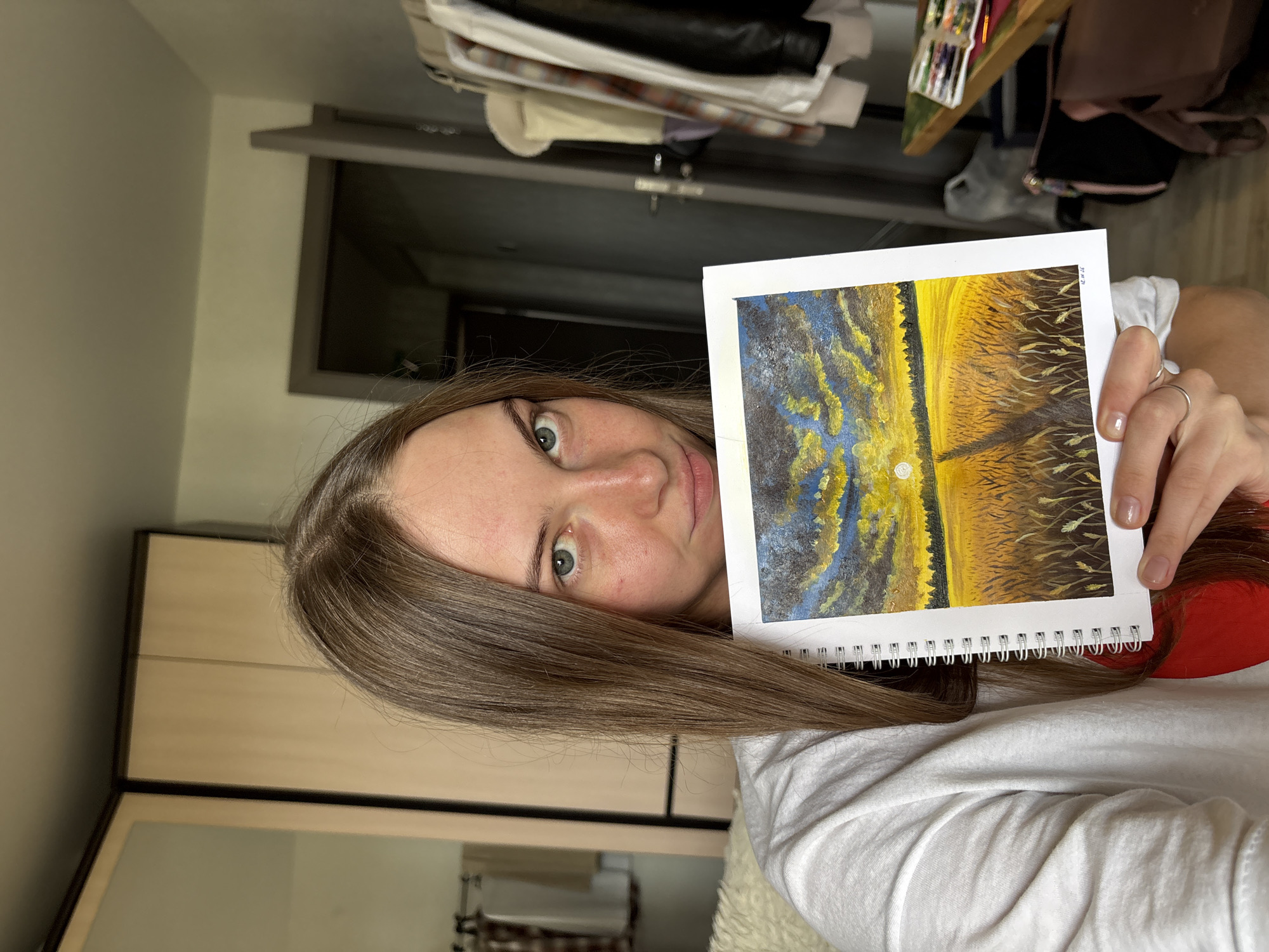 The image shows a young woman with long brown hair holding up a painting of a wheat field at sunset. She is wearing a white sweatshirt and has a red garment visible underneath. The painting depicts a path through a golden wheat field leading to a forest line under a dramatic sky with a setting sun. The background suggests an indoor setting with a closet, a doorway, and clothing on a rack. The woman's pose and the painting she holds indicate she is showcasing her artwork. The image does not contain any specific information or visual cues that would allow for its location to be identified within the city of Perm, Russia.