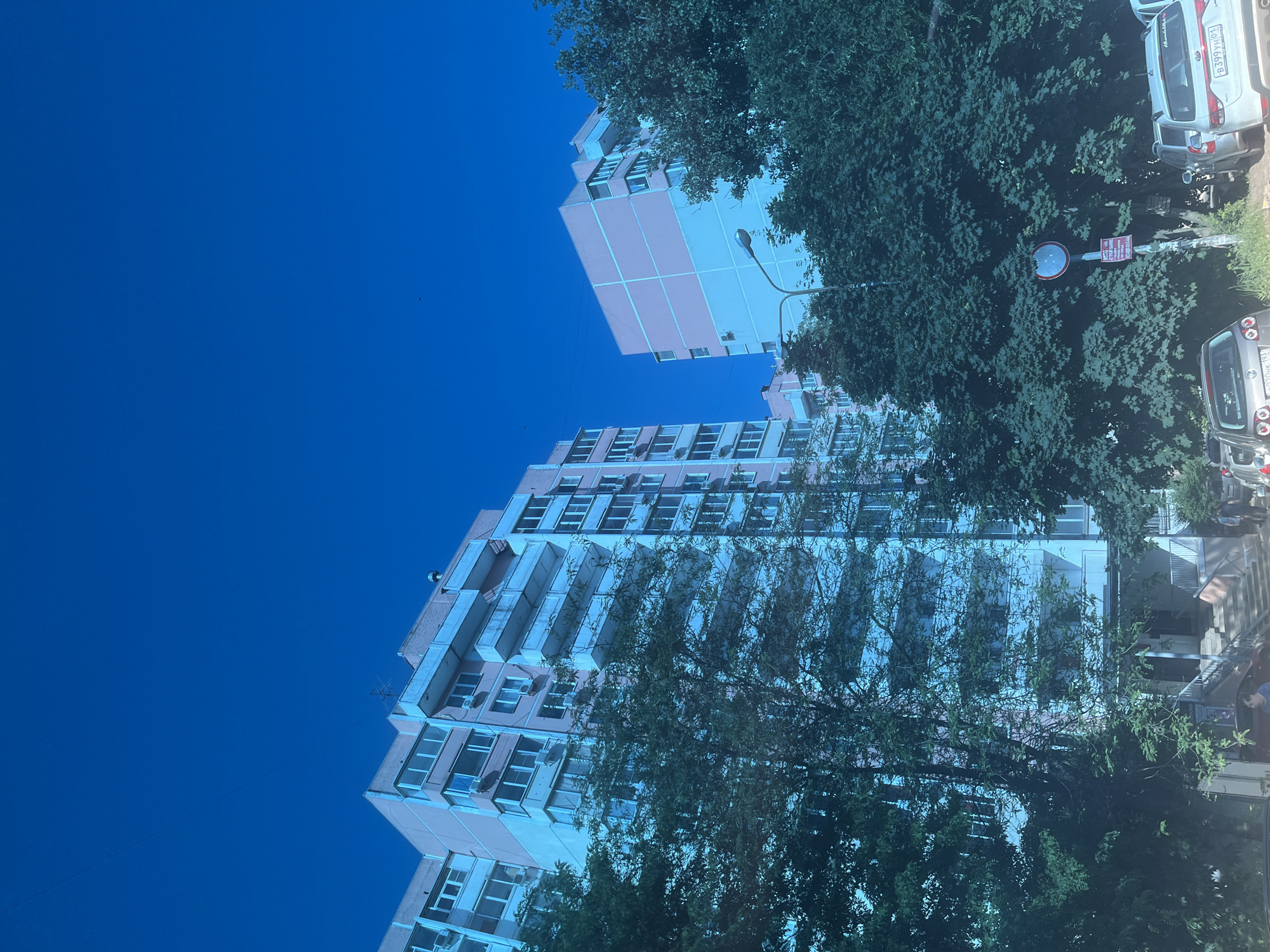 The image depicts a residential urban scene under a clear, deep blue sky. Two multi-story apartment buildings, predominantly pink/salmon with white or light blue balconies and window frames, are visible. The building on the left is more prominent, featuring multiple windows and several external air conditioning units. The building on the right shares a similar architectural style and color scheme.
Dense green deciduous trees occupy the mid-ground and foreground, partially obscuring the lower sections of both buildings and elements at ground level. A street light pole with a lamp is visible between the two buildings. Further to the right, behind the trees, a pole displays a circular prohibitive sign (white interior with a red border) and a smaller, rectangular red sign below it.
At ground level, several vehicles are present. A silver Kia Rio with Russian license plate B399YH 93 (Krasnodar Krai) is parked on the right. A grey car with a partial license plate P020HH 93 (Krasnodar Krai) is visible on the left. Steps leading to an entrance are visible on the left building.
Two individuals are partially visible: one near the entrance steps of the left building, and another near the silver Kia on the right. The scene is illuminated by bright daylight, characteristic of a summer day given the lush foliage.
Based on the visible license plates with the "93" region code, the location is within Krasnodar Krai, Russia. The architecture and urban setting indicate a residential area within the city of Krasnodar. The precise street address or specific building number within Krasnodar is not discernible from the provided imagery.