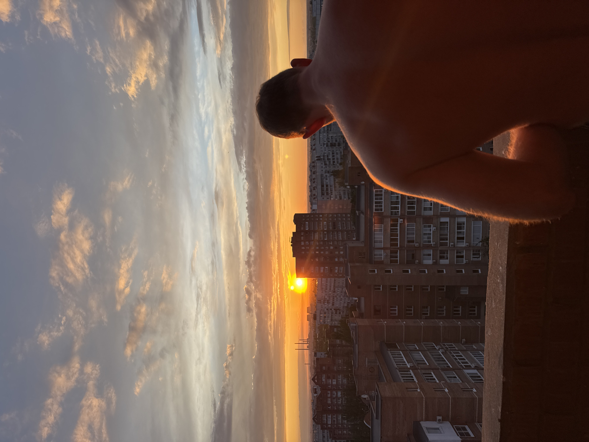 From an elevated urban perspective, likely a rooftop or high balcony, a shirtless individual is observed from behind, facing a setting sun. The person's head and upper back, illuminated by the warm, orange light of the sunset, show a clear outline and skin texture. A dark, concrete railing forms the immediate foreground barrier.
Beyond the railing, an expansive cityscape unfolds under the sunset. Numerous multi-story residential buildings, predominantly constructed from brick and concrete, populate the midground. Their facades display multiple windows and balconies, some with visible air conditioning units. The sun, a brilliant orange-yellow orb, is positioned above the horizon line, casting intense light across the scene and creating a strong lens flare effect in some areas. To the left of the sun, several tall industrial chimneys or smokestacks are discernible in the distant haze.
The sky is a prominent feature, transitioning from intense orange and yellow at the horizon to softer peach, pink, and pale blue-grey hues higher up. Scattered clouds, illuminated from below by the sun, display varying shades of orange, pink, and grey against the otherwise clear sky. The overall lighting is warm, with deep shadows in areas not directly touched by the sunlight. The location is an unknown city in an unknown country.