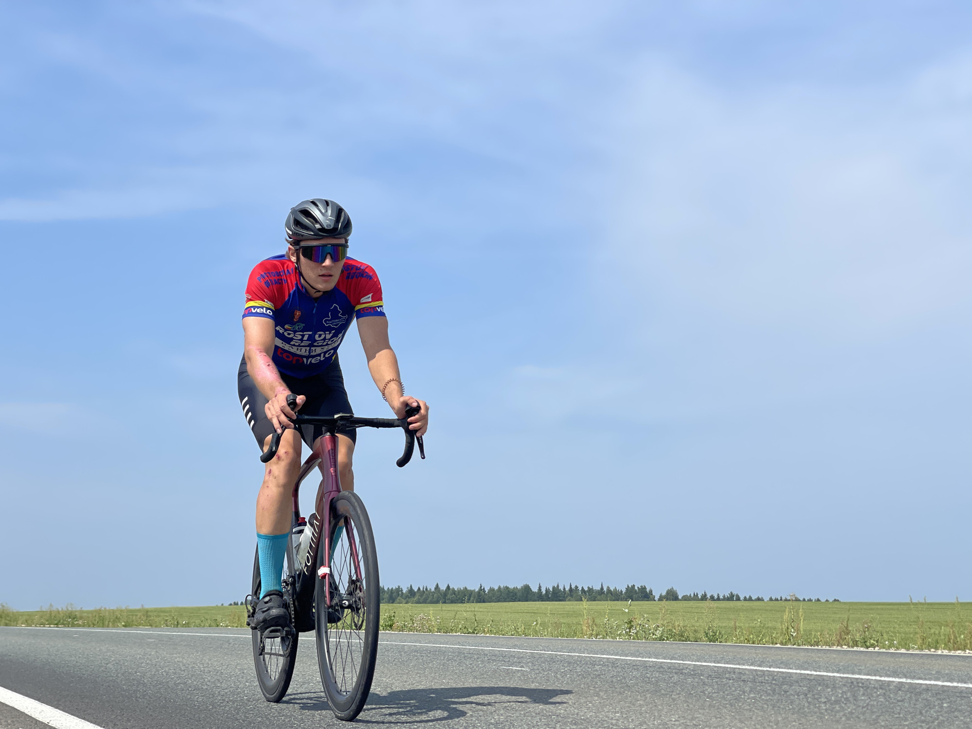 A male cyclist wearing a helmet, sunglasses, and a blue and red cycling jersey rides a red road bike on an asphalt road. He is wearing blue socks and black cycling shorts. The road is lined with green fields and a forest in the distance under a clear blue sky. The cyclist's legs appear to have scrapes and bruises. The image is taken in Niva, Russia.