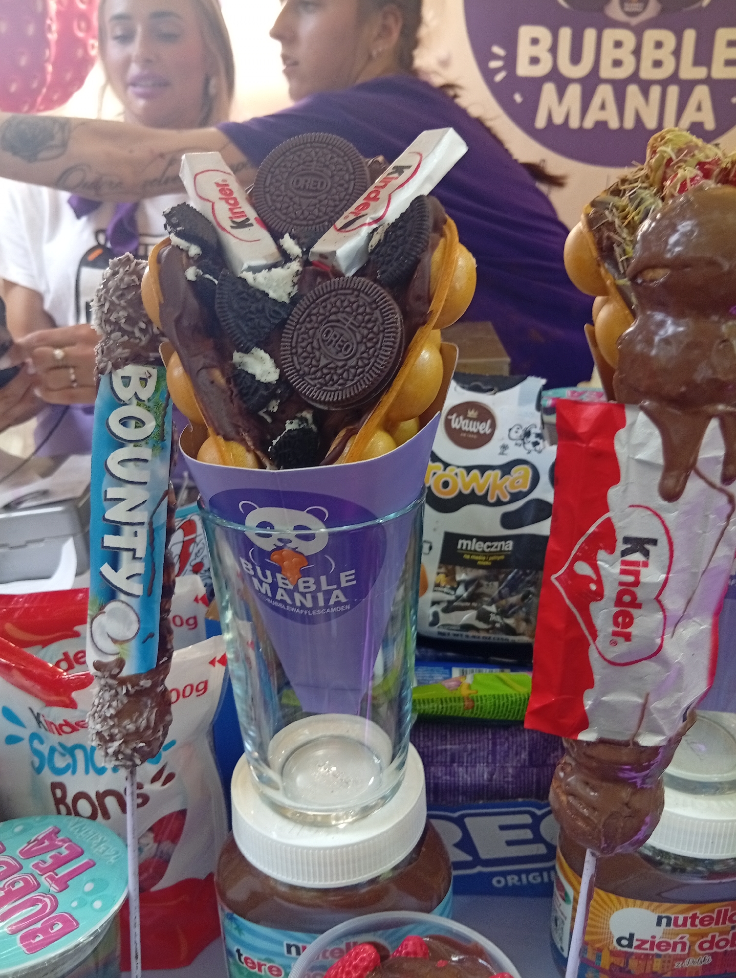 The image depicts a dessert stand, likely in Wrocław, Poland, specializing in bubble waffles and other sweet treats. In the foreground, there are several visually appealing desserts arranged on skewers. One prominent dessert features a bubble waffle base coated in chocolate and adorned with Oreo cookies and a Kinder chocolate bar. Another skewer displays a Bounty chocolate bar covered in coconut flakes and chocolate.
Behind the desserts, the background shows various branded packaging, including Kinder, Wawel, and Nutella, suggesting popular confectionery brands are used in the preparation of these items. A large purple cone with the "Bubble Mania" logo is visible, reinforcing the establishment's theme. Two women are partially visible in the background, with one wearing a purple top and the other a white top and a tattoo on her arm. The overall impression is that of a vibrant and enticing dessert stall ready to serve customers.