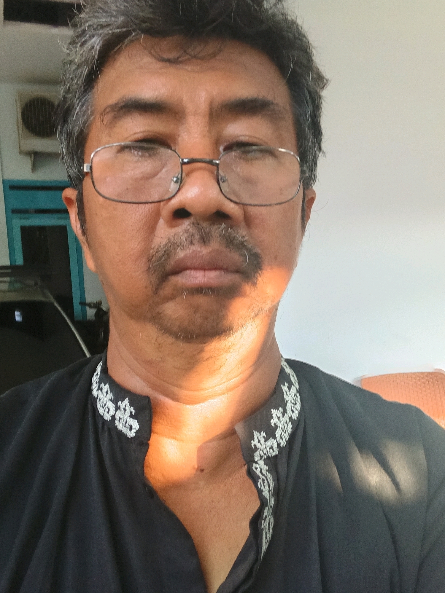 A close-up, selfie-style photograph features an East Asian man with graying dark hair, wearing glasses, a dark shirt with white embroidery around the collar, and a mustache. He is outdoors, with a white wall and a blue-framed window visible in the background, suggesting a residential setting. A car is partially visible to the left, and an air conditioning unit is mounted on the wall. The lighting indicates it is daytime, with sunlight casting shadows on the man's face and shirt. The image appears to have been taken in Tanggul Wetan, Indonesia.