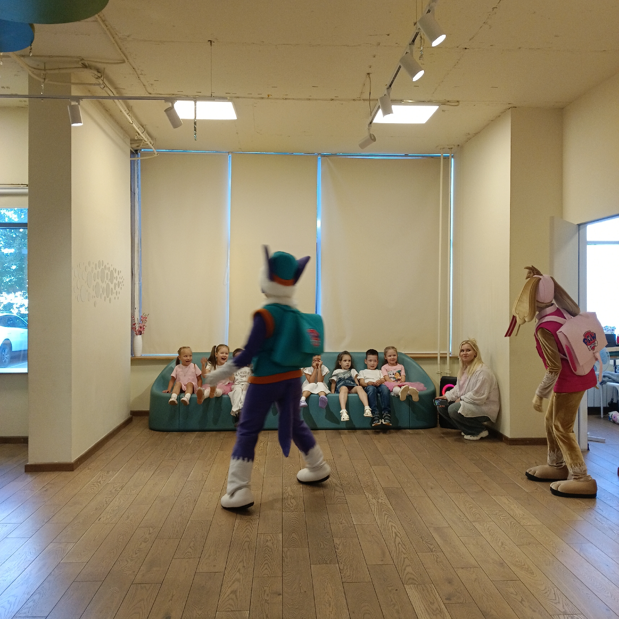 In an indoor studio or event space with a light-colored wooden floor, two costumed characters are present. On the left, a character dressed as a blue and teal cat-like figure with purple pants and white boots stands facing a group of children seated on a teal couch. The children, ranging in age from roughly 5 to 8 years old, are looking towards the costumed figure. On the right side of the frame, another costumed character, resembling a rabbit with large floppy ears and a pink backpack, is partially visible and facing towards the right edge of the image. A woman in casual clothing is seated on the floor near the rabbit character, holding a mobile phone. The environment features large windows with cream-colored blinds, ceiling lights, and a structural pillar. The overall scene depicts a children's event or performance.