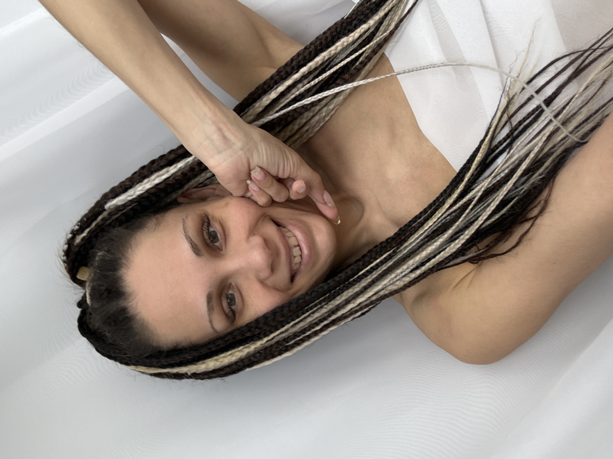 A young woman with light skin, brown hair in black, brown, and blonde braids, and green eyes, is lying on a white surface with a white cloth partially covering her chest. She is smiling and looking directly at the camera. One of her hands is raised near her face. The location is likely indoors but details to confirm this with certainty is lacking. No information to confirm this is Rusanovo, Russia.
