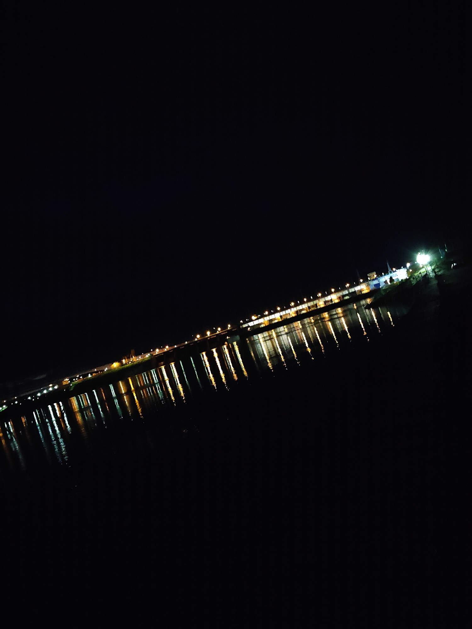 The image displays a nighttime scene in Chaikovsky, Russia, focusing on a long, illuminated structure stretching horizontally across a calm body of water, likely the Kama River at the Votkinsk Dam. The structure is adorned with numerous artificial lights, predominantly warm yellow-orange and some white, which are evenly spaced along its length. These lights create distinct, elongated, vertical reflections on the dark water surface below, indicating a smooth water current. The sky above is completely dark, providing no discernible features. On the far right, a cluster of brighter lights suggests a more prominent section of the structure or an adjacent landmass. No individuals, vehicles, or specific activities are visible within the frame.