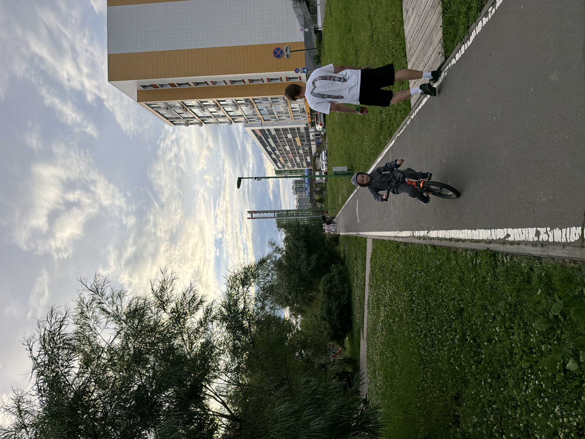 In Kazan, Russia, a young child in a grey hooded jumpsuit rides a small orange bicycle on an asphalt path lined with white markings. To the right of the path, a man wearing a white t-shirt with a graphic design on the back and black shorts walks away from the viewer. A lush green lawn with scattered small white flowers borders the path on the left and right. In the background, modern apartment buildings with beige and white facades stand tall under a cloudy sky. Trees and other vegetation are visible on the left side of the image, with distant construction cranes and more residential buildings in the far background. The scene depicts an outdoor recreational area within a residential urban environment.