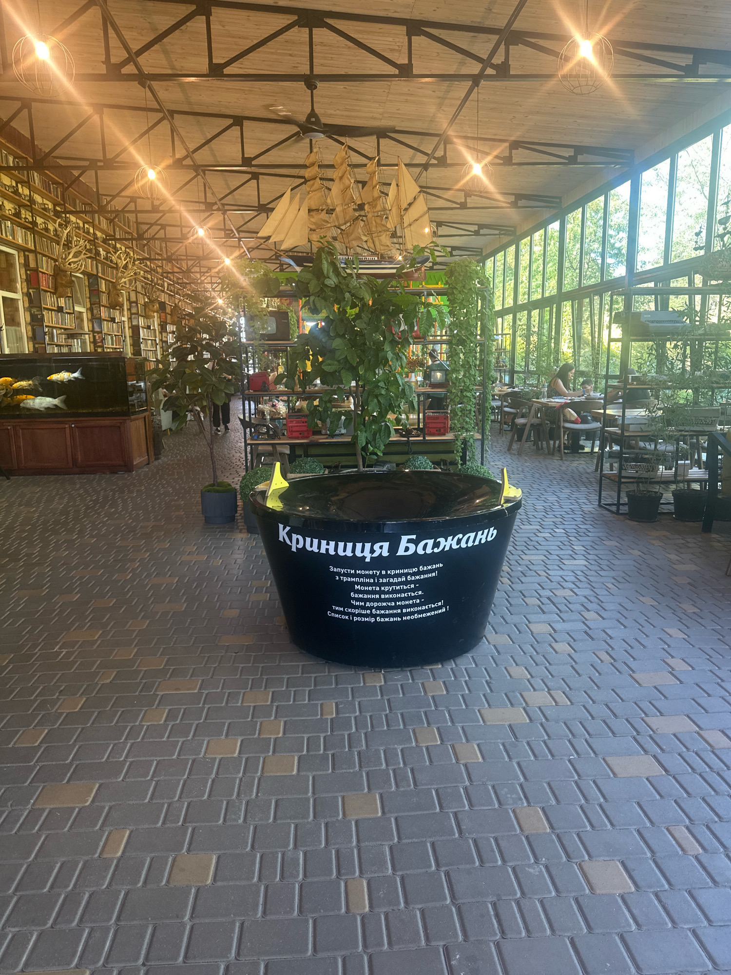 This is a full shot of the interior of a cafe in Molodizhne, Ukraine, possibly an all-season terrace due to the large windows and exposed beams. The flooring is made of interlocking brick pavers in shades of gray and tan. In the foreground, a large black tub, labeled in Ukrainian "Криниця Бажань" (Well of Wishes), is filled with water and has a yellow object floating in it. Plants are placed around the tub. To the left, a large aquarium with koi fish is visible. In the background, shelves stocked with books line the wall, and decorative ship models hang from the ceiling. The space is illuminated by industrial-style pendant lights and natural light streaming through the windows, which offer views of greenery outside. Several tables and chairs are set up for dining, and a few patrons are seated and interacting. A ceiling fan is also visible.