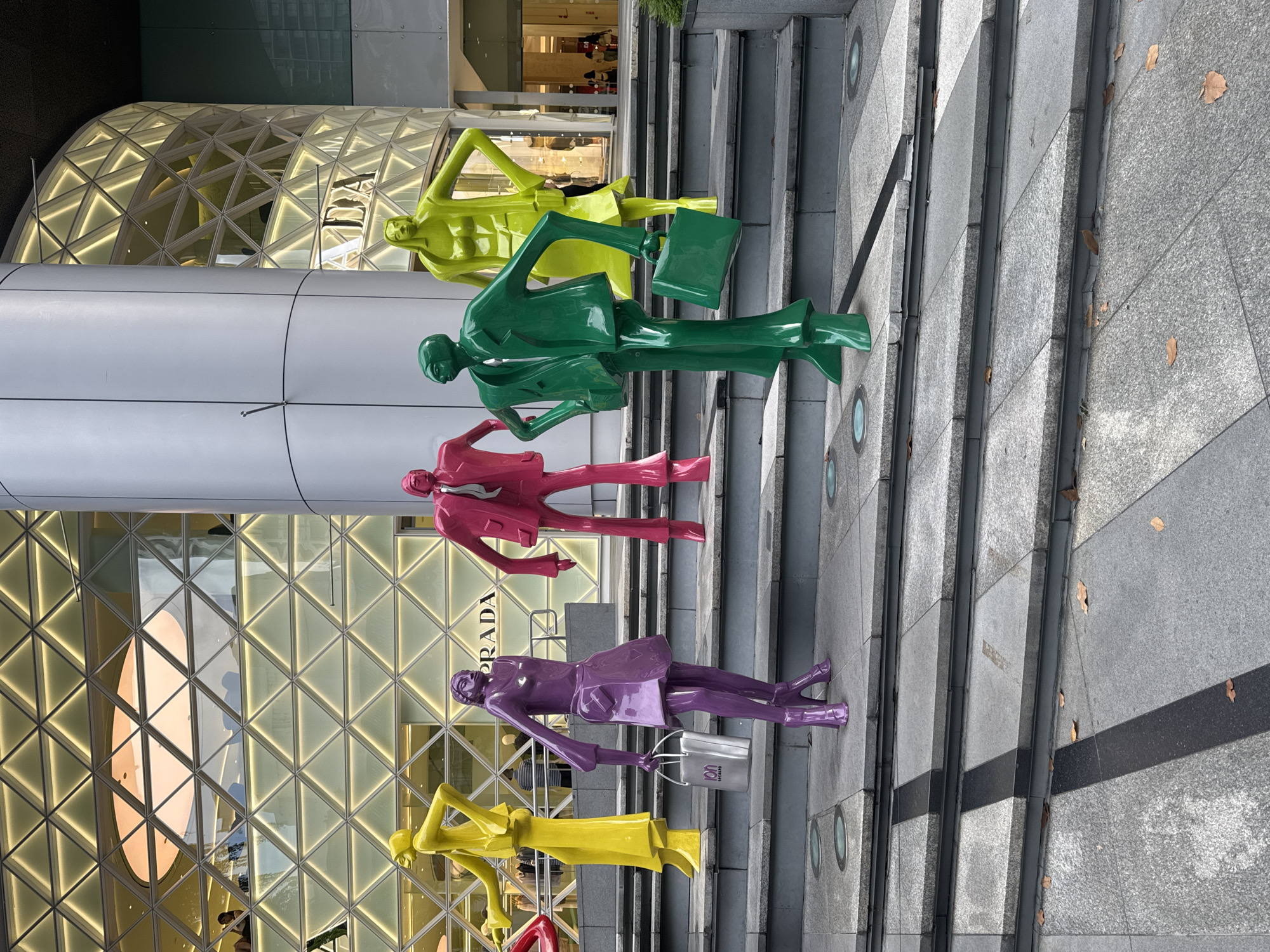 This image depicts a public art installation featuring five brightly colored, stylized mannequin-like sculptures in front of the ION Orchard mall in Singapore. The sculptures, colored yellow, purple, pink, green, and lime green, are positioned on a wide set of granite stairs leading up to the entrance of the modern building, which features a distinctive geometric facade with integrated lighting. The yellow sculpture is on the far left, followed by the purple one holding a silver shopping bag with the "ION ORCHARD" logo. Next are the pink and green figures, with the lime green sculpture on the far right. The background shows the illuminated facade of the mall, with a glimpse of shoppers inside through the glass entrance. The scene is set outdoors on a bright day, with a few fallen leaves scattered on the stairs and ground.