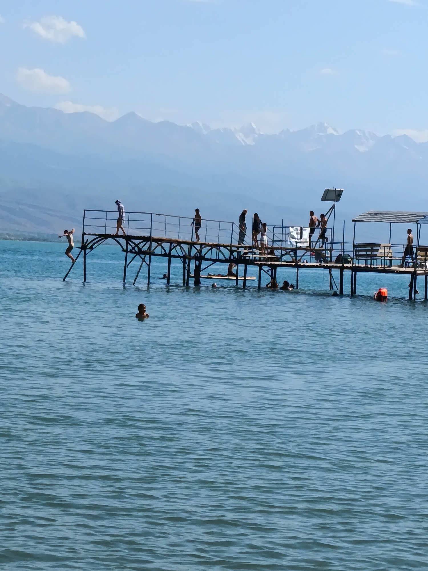 The image depicts a pier on a body of water, likely a lake or reservoir, with a mountainous backdrop. The pier is constructed of wood and supported by metal stilts, with a railing along its length. Several individuals are visible on the pier and in the water. On the pier, people are standing and walking, and one person appears to be in the process of jumping into the water. In the water, some individuals are swimming, with one person visible near the surface and another near an orange buoy. The water is a calm, blue color, and the sky is clear with some scattered clouds. The mountains in the background are snow-capped, suggesting a high-altitude location. The overall scene conveys a sense of leisure and recreation.