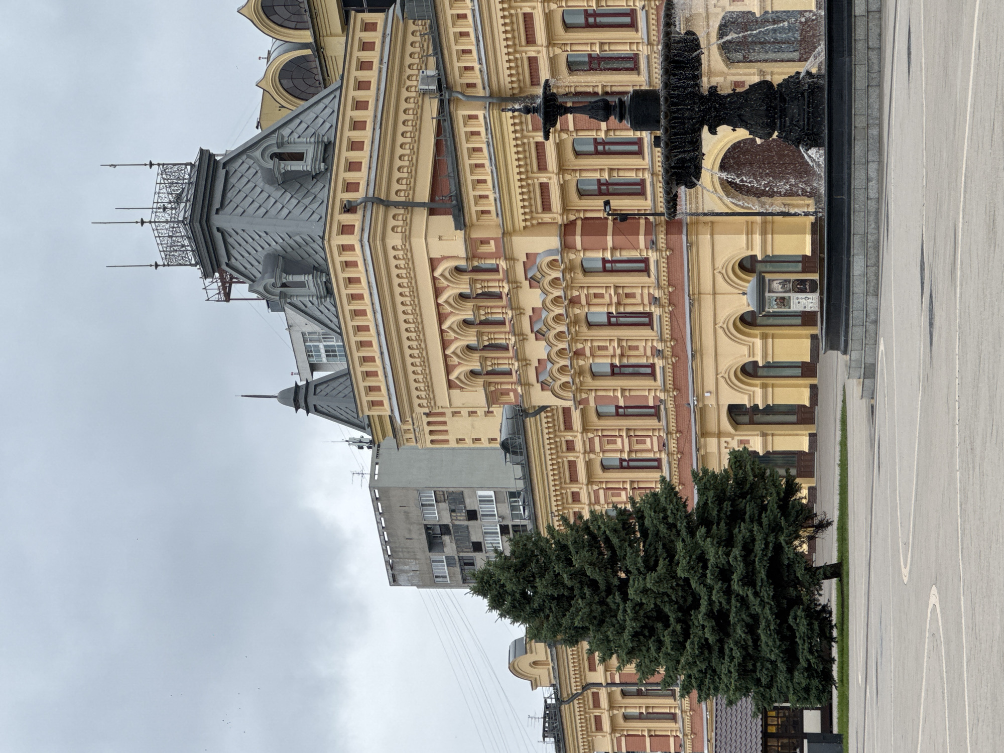 The image shows a view of the Nizhny Novgorod State Academic Opera and Ballet Theatre, located in Nizhny Novgorod, Russia. The theatre's ornate yellow facade with red trim is a prominent feature, characterized by arched windows and decorative architectural elements. To the left of the theatre, a large, dark green evergreen tree stands in a paved plaza. In the foreground, a multi-tiered black fountain is active, with water cascading from its upper levels. In the background, a utilitarian, gray apartment building with many windows stands in contrast to the historical architecture of the theatre. The sky is overcast and gray.