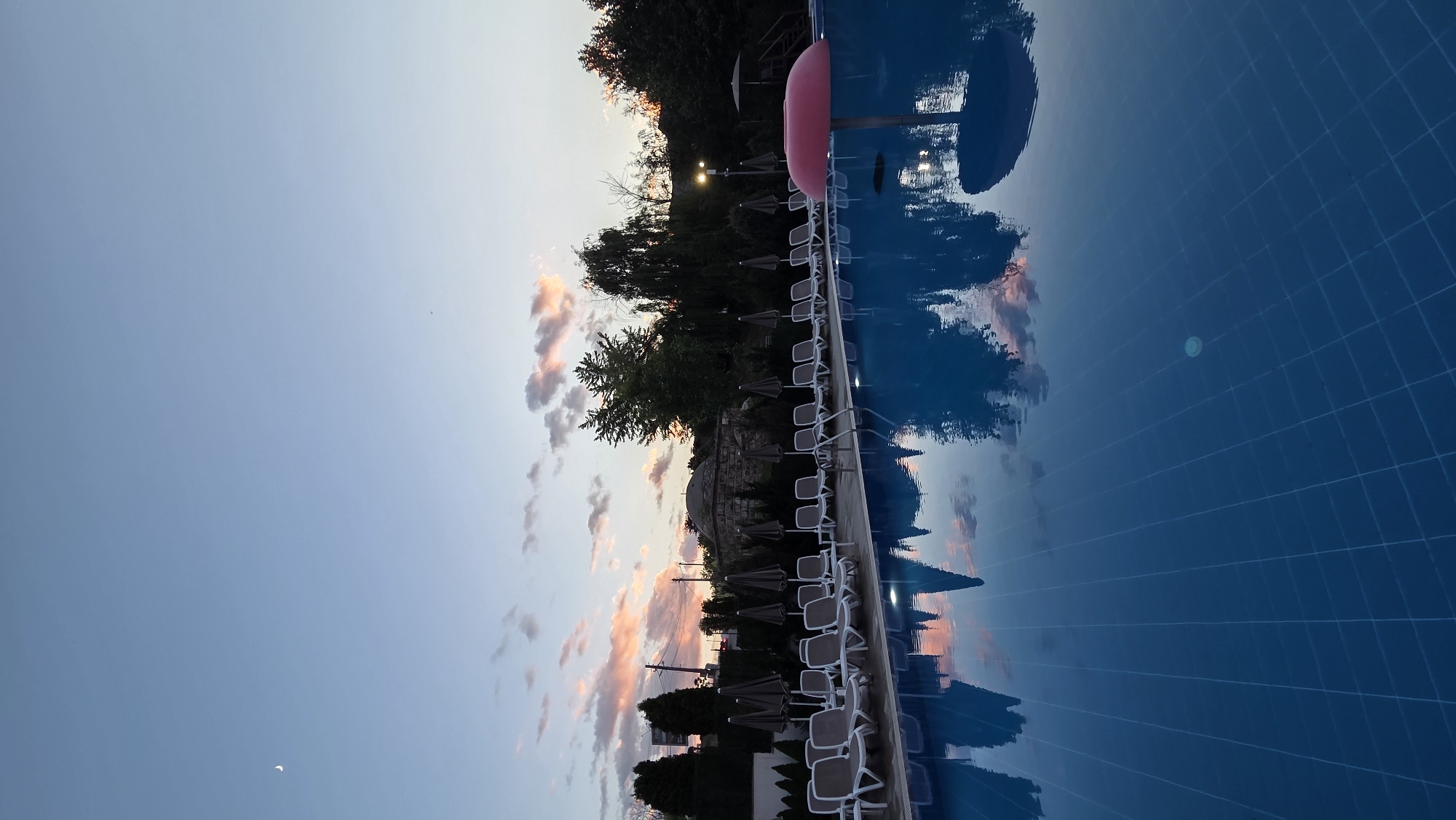 An outdoor swimming pool is shown at dusk, with a clear sky exhibiting a faint crescent moon and soft pink clouds. The pool water is a vibrant blue and reflects the sky, clouds, and surrounding trees and lounge chairs. A row of empty lounge chairs with grey umbrellas is lined up along the pool's edge. In the water, a pink, mushroom-shaped pool float is visible. In the background, there are trees, a building with a dome, and streetlights. The scene appears serene and is situated in Bania, Bulgaria.