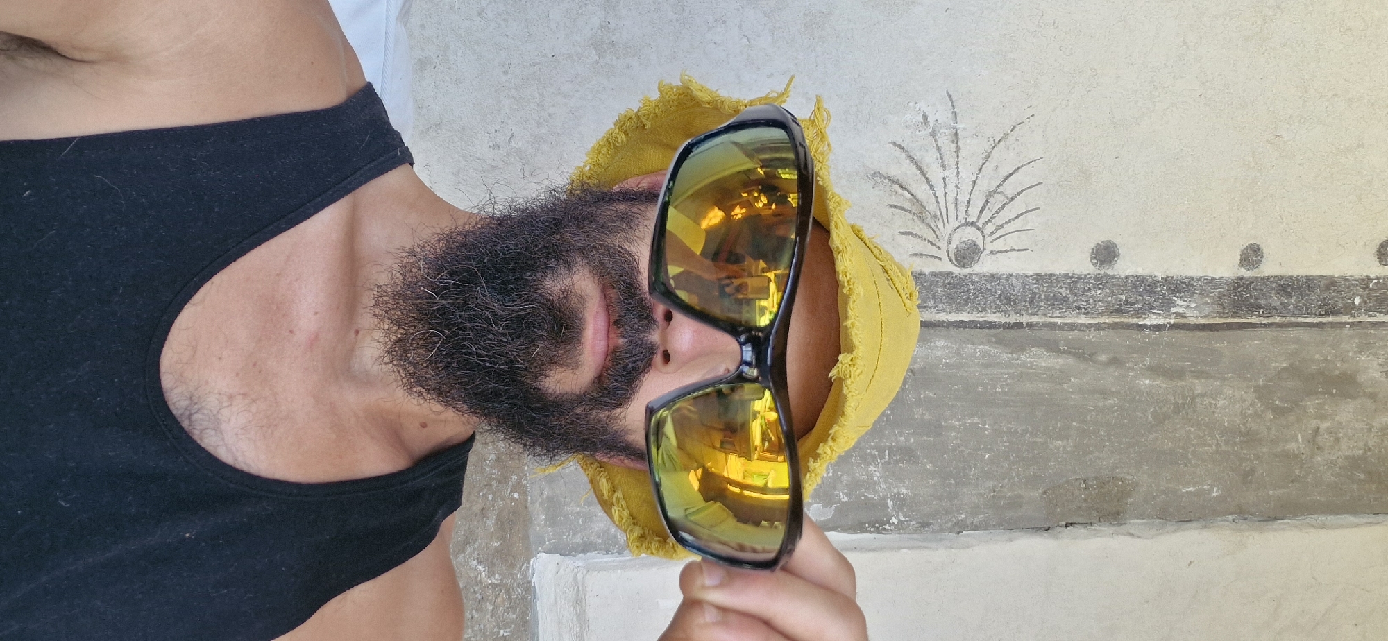 A man with a beard and a yellow hat is holding up sunglasses to his face. The sunglasses have yellow, reflective lenses that show a distorted reflection of lights and possibly a ceiling. The man is wearing a black tank top. He is standing in front of a rough, textured wall with some faint drawings or markings on it. There is a hint of a white surface to his left. The background wall appears to be a light color, possibly stucco, with a darker vertical stripe and some circular, dark marks. The drawing on the wall is abstract, resembling an eye with radiating lines. The overall scene appears to be outdoors, possibly in a courtyard or under an overhang, given the natural lighting. The location is the city of Wattenwil, Switzerland.