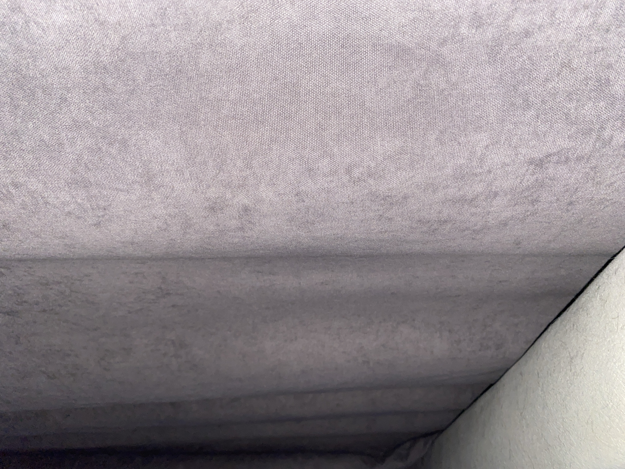 The image displays a close-up, static view of interior fabric surfaces. The primary element is a textured, light gray or muted purple-gray woven fabric that covers most of the frame. This material appears to have a fine, slightly plush nap, characteristic of automotive headliners or upholstery fabric.
A horizontal fold or crease extends across the middle of the image, separating a brighter, upper section from a darker, shadowed lower section of the same fabric. The lower portion shows subtle undulations and appears darker due to diminished lighting.
On the right side, a sharp diagonal line indicates the edge of a different interior component. This contrasting surface is off-white or very light gray and exhibits a distinct, possibly softer or fuzzier, texture compared to the main gray fabric.
The lighting is dim and indirect, contributing to soft shadows and emphasizing the tactile qualities of the materials. No people, objects (other than the fabric itself), or activities are visible. The scene suggests an interior environment, likely a vehicle or a room with upholstered elements. There is no visible text.