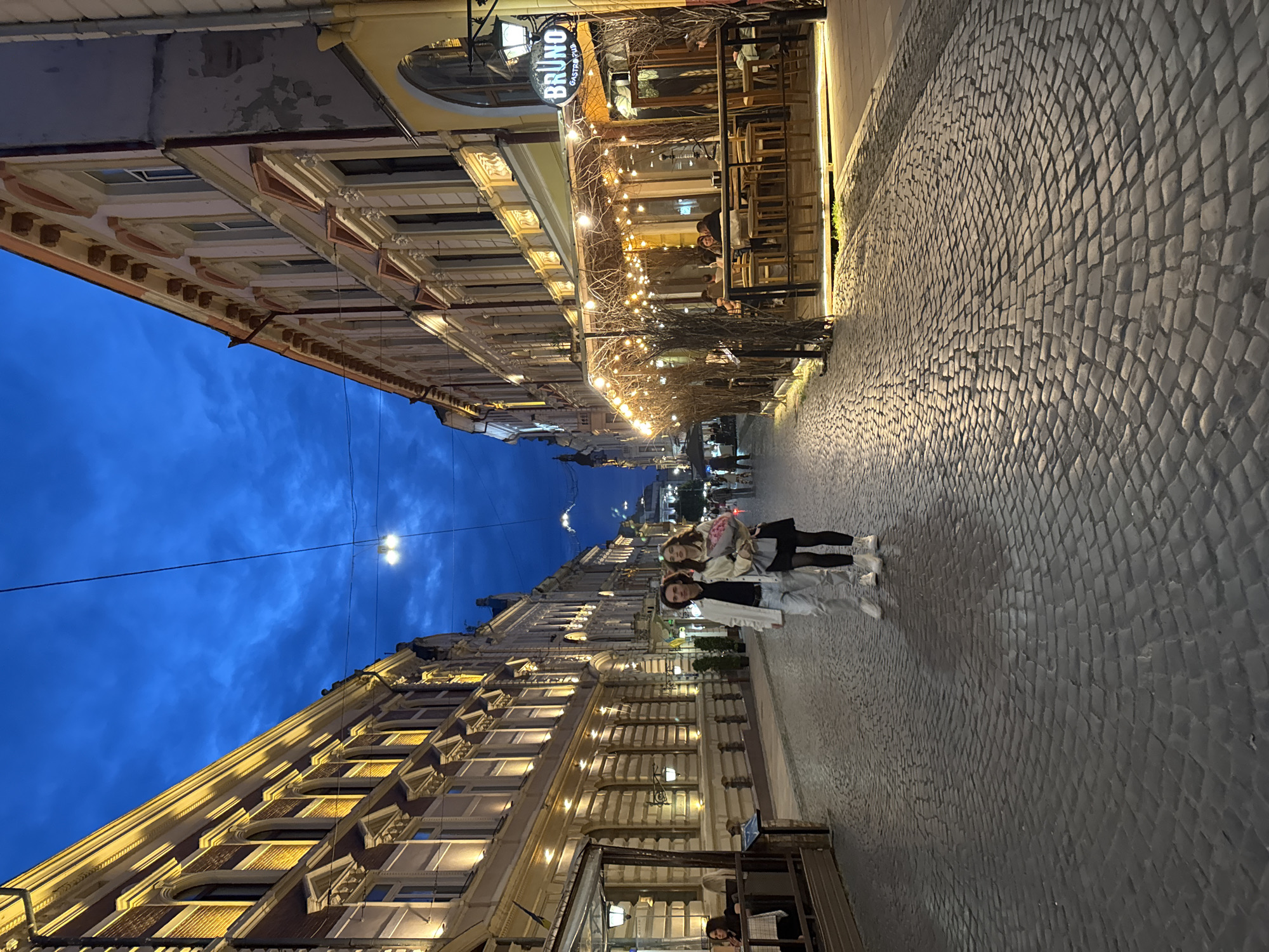The image depicts a cobblestone street in Chernivtsi, Ukraine, at dusk or early night. The sky is a deep blue, with some clouds visible. The street is lined with ornate, historic buildings on both sides, illuminated by warm, artificial light. The building on the right features a restaurant with outdoor seating, adorned with string lights and bare branches. A sign for "BRUNO Gastro Pub" is visible.
In the foreground, a young couple is walking down the street. The man is wearing a white jacket, a black shirt, and light-colored pants. The woman is wearing a black dress and white boots, and she is carrying a bouquet of pink flowers. They are walking towards the viewer. Further down the street, other people can be seen dining at the outdoor tables of the restaurant and walking along the sidewalk. The cobblestone pavement shows a circular pattern in the foreground, suggesting the street is well-maintained and potentially a pedestrian area. The overall atmosphere is calm and romantic, with the lighting creating a cozy ambiance.