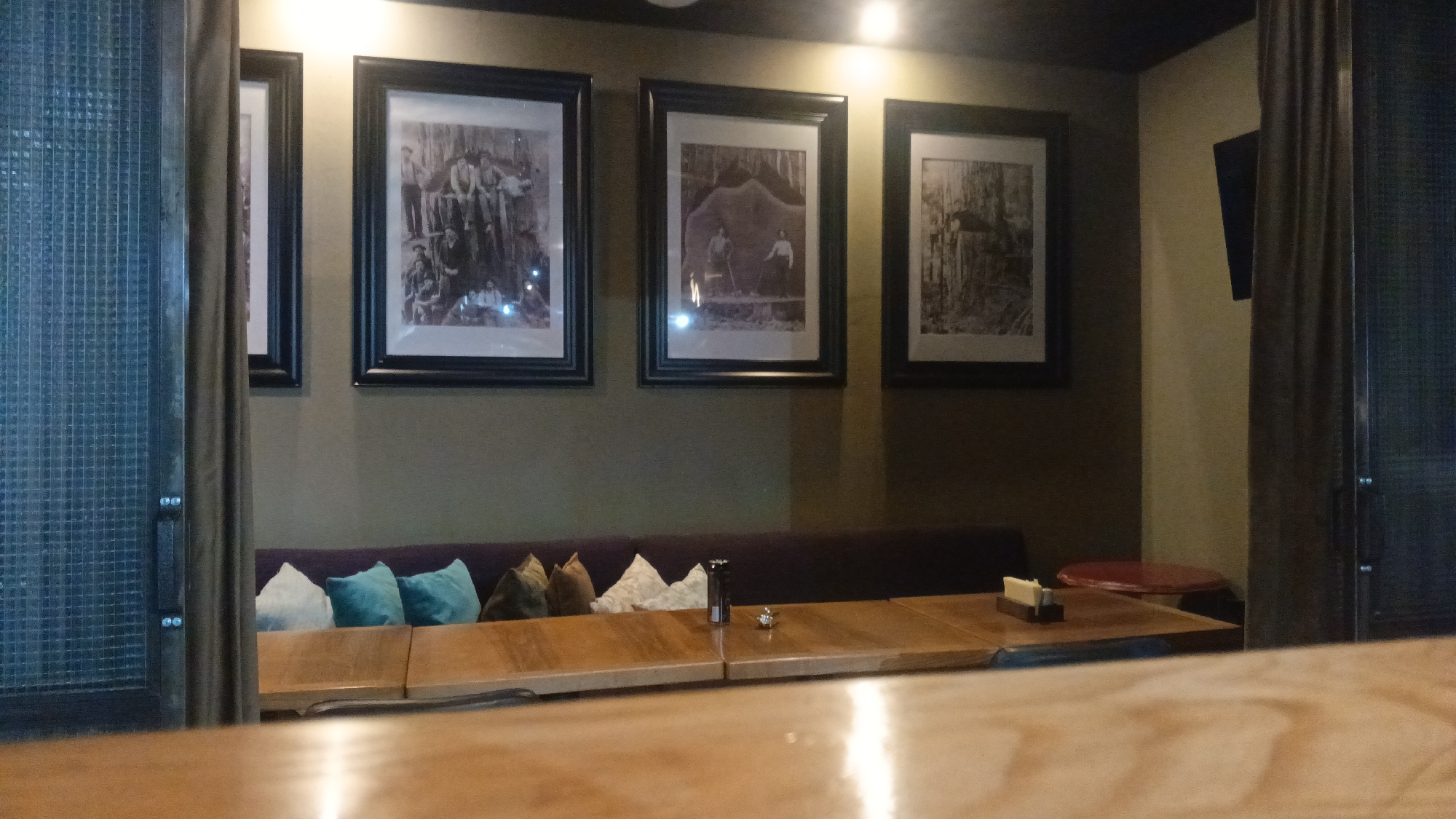 The image depicts an indoor scene, likely a restaurant or cafe, with a focus on a seating area and decorative elements. On the wall, three framed black-and-white photographs are displayed. The leftmost photograph appears to show a group of people in historical attire, possibly a family or workers. The central photograph features two men in a wooded setting, one holding an axe. The rightmost photograph, partially obscured by a curtain, also seems to depict a scene in nature.
In front of the wall, a long, dark-colored banquette seat is adorned with several decorative cushions in varying colors, including white, teal, and brown. A wooden table is positioned in front of the banquette, with several sections indicating it is a modular or extendable table. On the table, there are a few small objects, including what appears to be a salt and pepper shaker set and a small card holder. The foreground shows another wooden table surface, which is out of focus and reflects light, suggesting it is closer to the camera.
To the left of the seating area, a glass partition with a metal grid pattern is visible, suggesting an entrance or an architectural feature. A curtain hangs on the right side of the frame, partially covering a wall-mounted screen or television and a red circular table in the background.
The lighting in the scene is dim, with warm, overhead lights illuminating the wall and the photographs. The overall ambiance suggests a cozy and perhaps vintage-inspired interior.
The location is Mytishchi, Russia.
