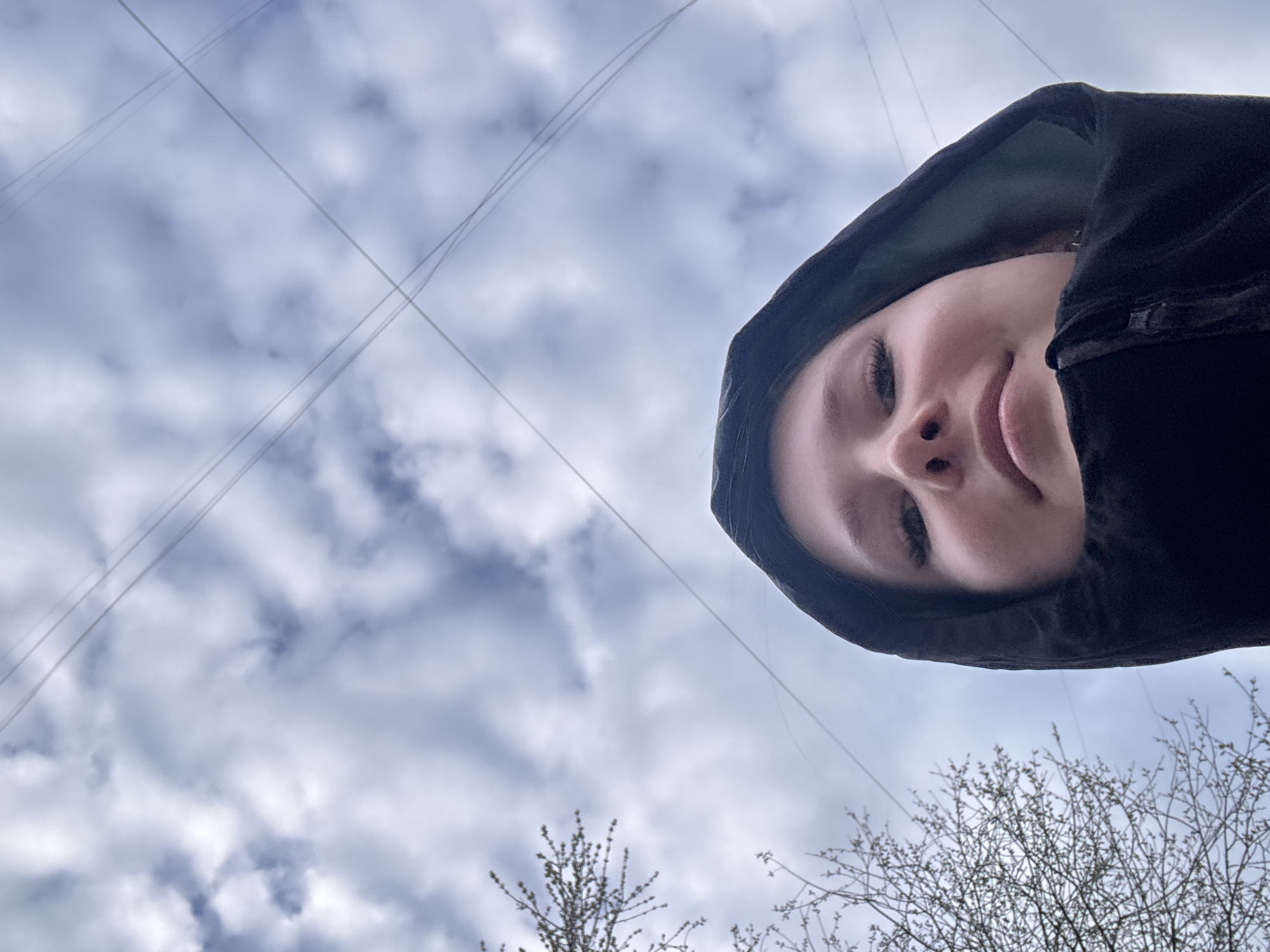 A low-angle photograph shows a person wearing a black hooded jacket, their face the most prominent feature, set against a cloudy sky. Electrical wires are visible stretching across the sky, and bare tree branches are present in the lower part of the frame. The location is inferred to be the city of Chaikovsky, Russia.