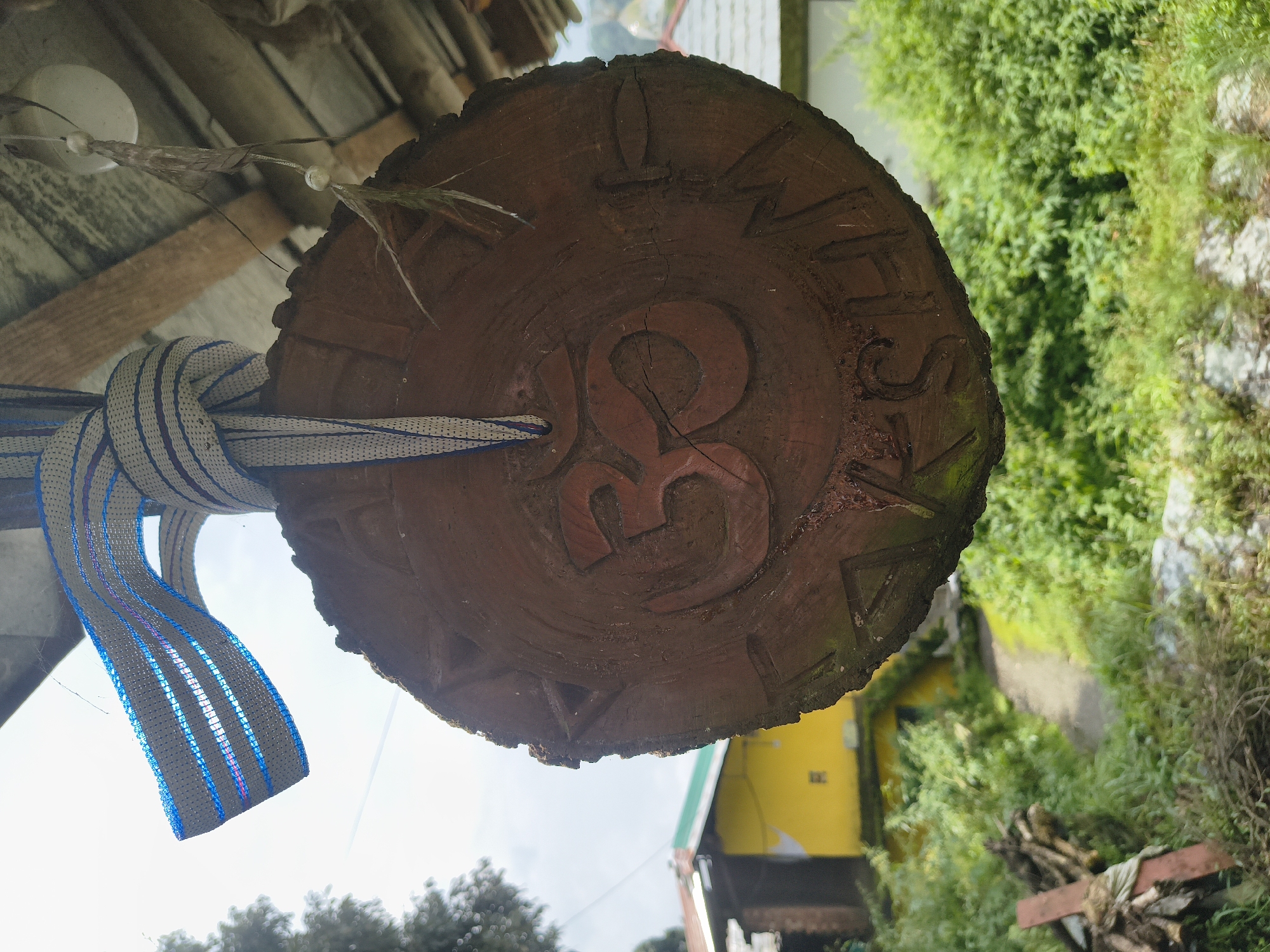 A circular wooden carving of the Om symbol hangs from a woven blue and white strap tied to an overhang. The Om symbol is centrally carved into the wood, with additional carved symbols or text around the perimeter. The carving is brown and appears weathered, with moss or lichen visible on the edges. In the background, a yellow building with a green roof is visible, along with lush green vegetation and a dirt path. The scene suggests a rural or semi-rural setting, possibly in a mountainous region given the cloudy sky and the style of the building. The specific location is Haini, India. There are no people visible in the image.