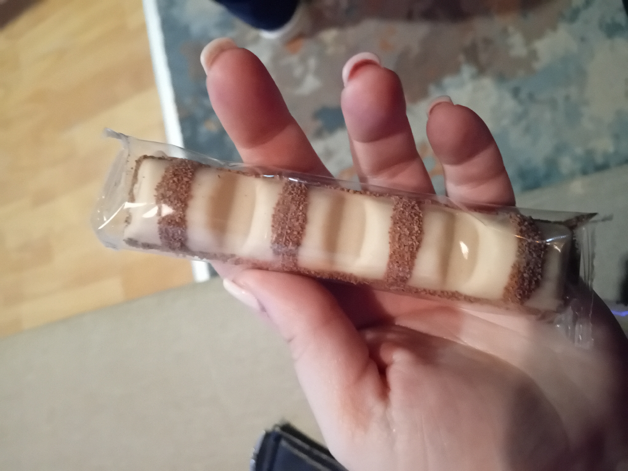 A hand holds a Kinder Bueno White chocolate bar in its plastic wrapper. The chocolate bar is white with brown sprinkles. The background is blurred but appears to be an indoor setting with a wooden floor. The location is Unknown, Unknown.