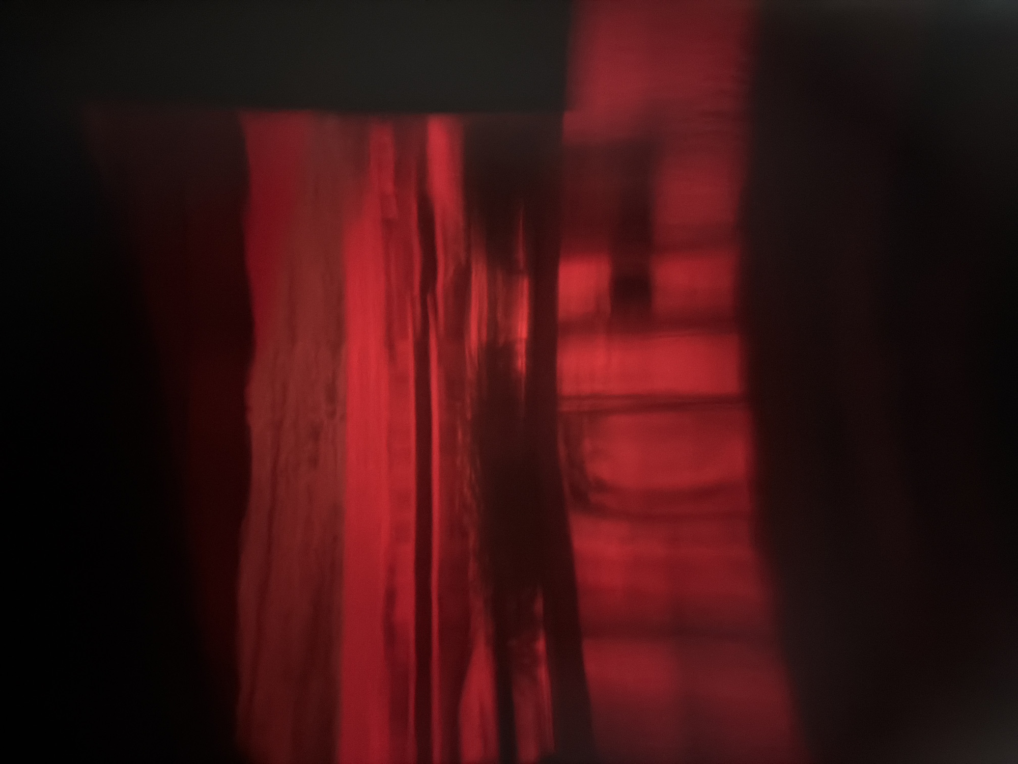 The image shows a dimly lit interior with red lighting. There appear to be vertical elements, possibly fabric or curtains, and what seems to be a window or opening with some undefined shapes. The overall scene is obscured and blurred, making it difficult to discern specific details. No specific location in Budva, Montenegro, can be determined from the image.
