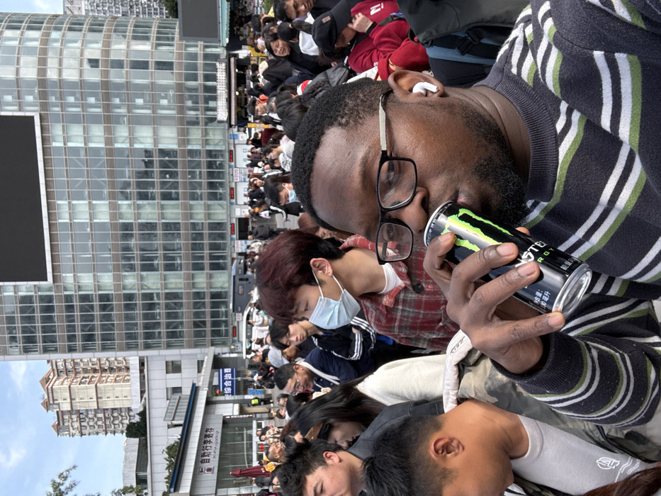An outdoor urban scene in Jiulongpo District, China, features a dense crowd and architectural structures under a partly cloudy sky. In the foreground, a Black male with a beard, wearing black-framed glasses and a dark striped sweater, is depicted drinking from a black and green Monster Energy beverage can. A white wireless earbud is present in his right ear. Behind him, a large group of predominantly East Asian individuals occupies the midground. One East Asian male with reddish-brown hair is visible to the left of the foreground subject, wearing a red plaid shirt and a light blue medical mask.
The background is dominated by a multi-story modern building featuring a reflective glass facade and a prominent dark screen on its upper portion. To the left of this structure, a lower grey concrete building displays Chinese characters translating to "Self-service luggage storage." Further left and behind, a residential building with light-colored walls and reddish-brown roof accents is partially visible. The scene suggests a busy public area.