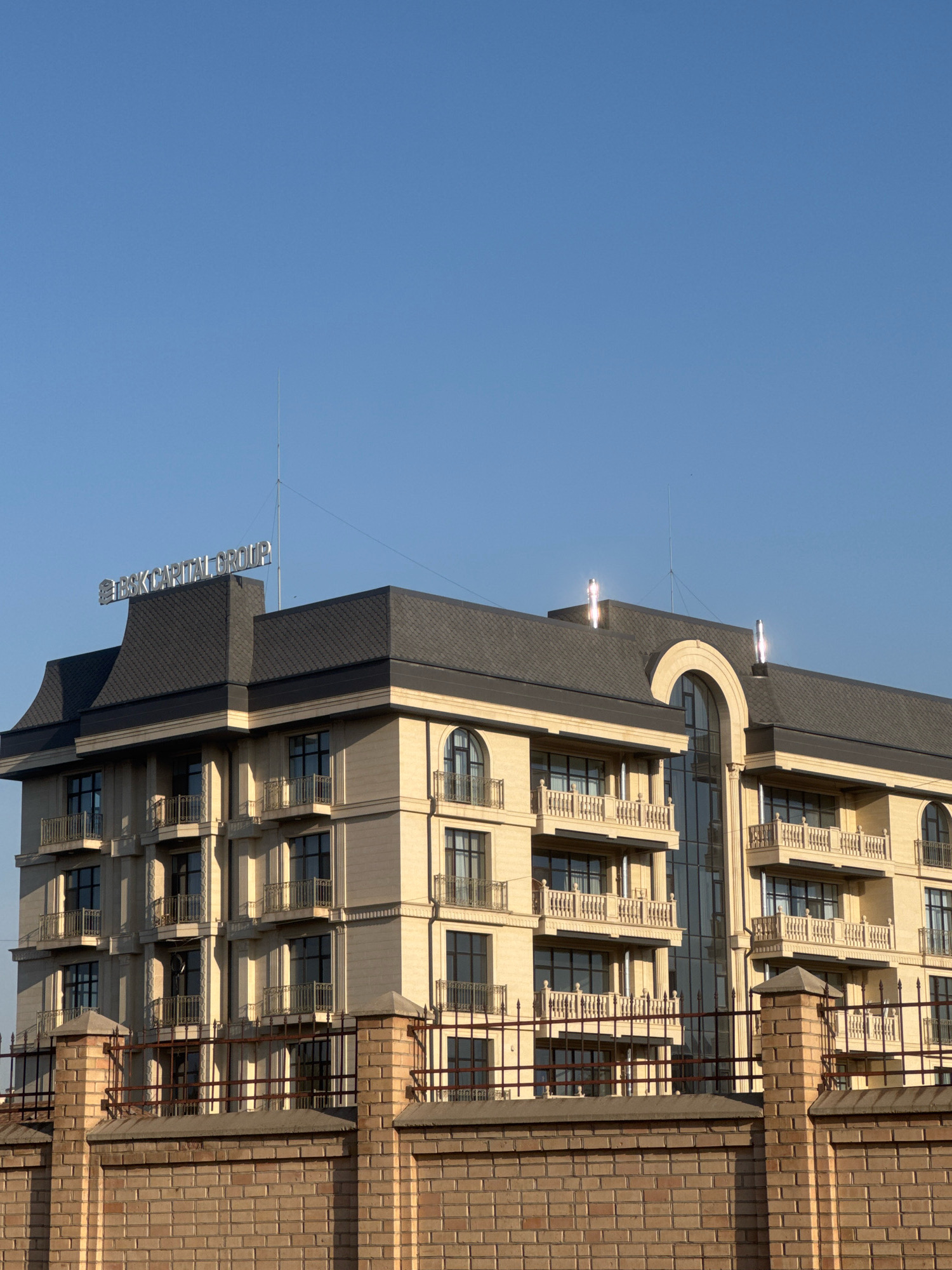 The image shows a modern, light-colored multi-story building with a dark gray roof, situated in Bishkek, Kyrgyzstan. The building features numerous windows and balconies with ornate railings. A sign reading "BSK CAPITAL GROUP" is visible on the roof. A brick wall topped with a metal fence is in the foreground, partially obscuring the lower levels of the building. The sky is clear and blue. There are no people visible in the image.