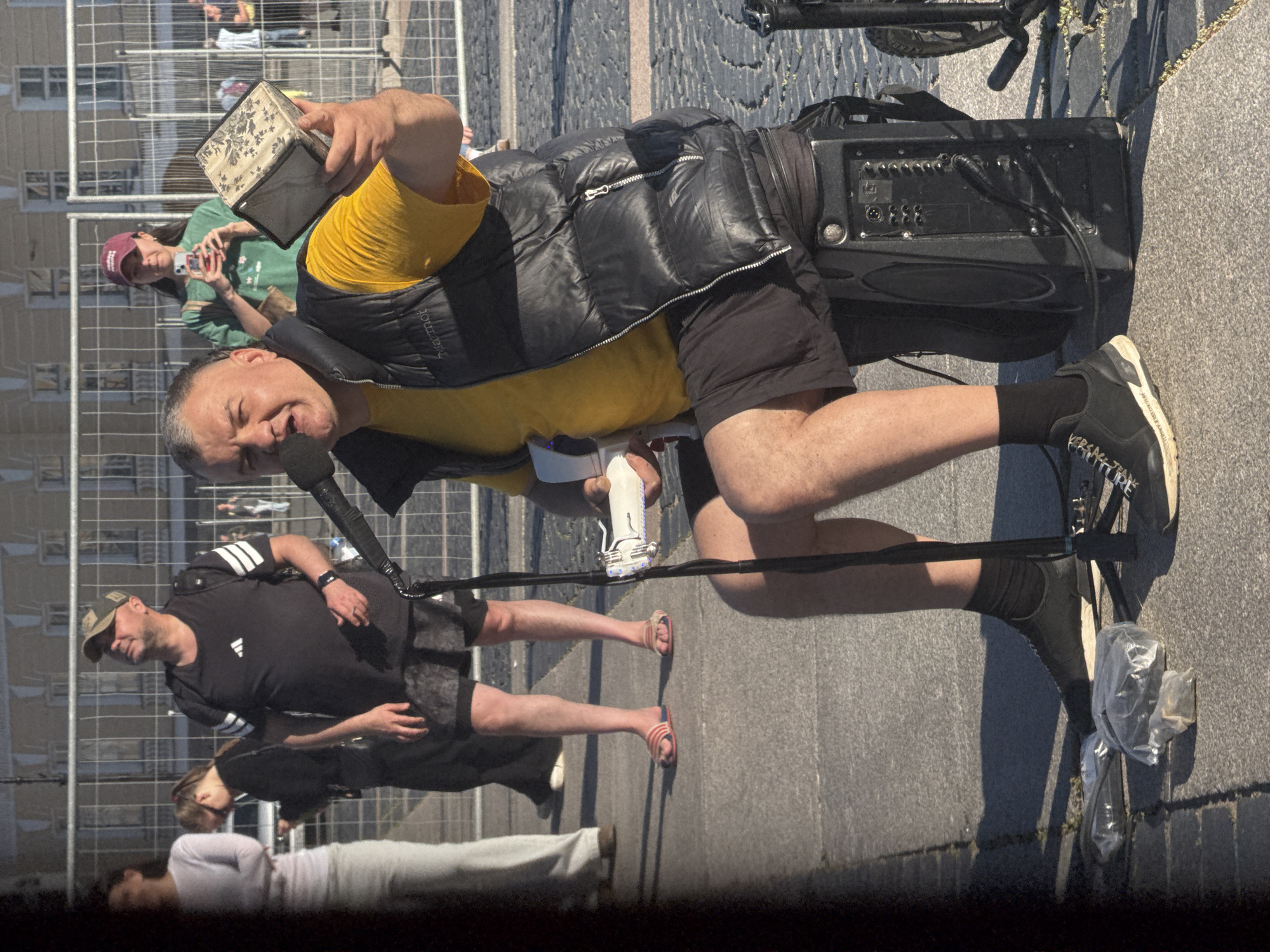 The image depicts a man performing music on a street in Saint Petersburg, Russia, near a fence. He is seated on a black amplifier, wearing a yellow t-shirt, a black puffer vest, black shorts, black socks, and black sneakers. He holds a microphone in his right hand and a decorative object in his left hand. A black cable runs from the microphone stand to the amplifier. To his left, on the pavement, are several plastic bags. In the background, a fence separates the performer from a street scene with other people. One woman in the background is holding a phone and appears to be taking a picture. The ground is paved with stone.