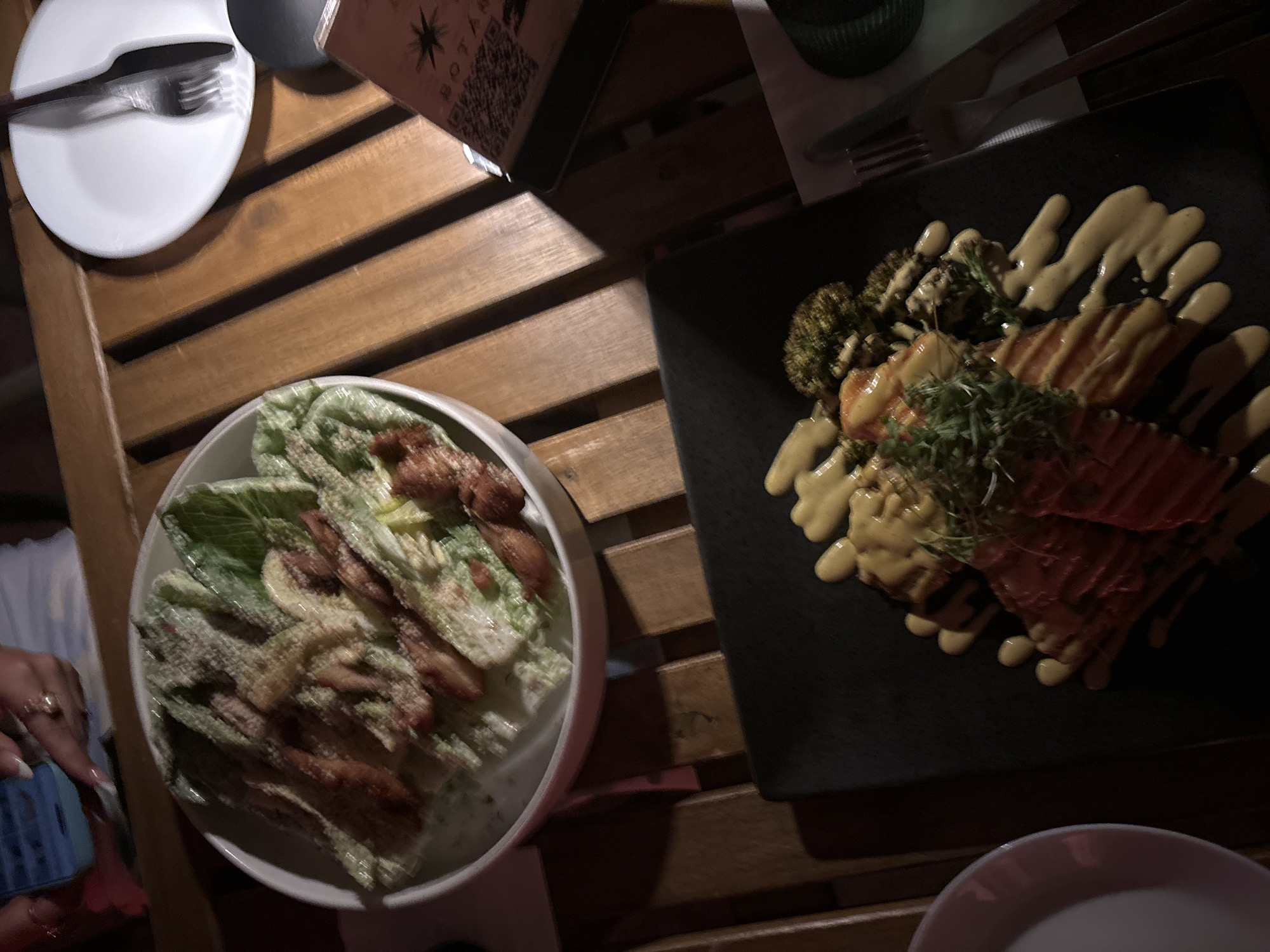 This media file captures a close-up, overhead view of a dining experience on a wooden table, likely at a restaurant in San Juan, USA. The scene features two dishes and tableware.
On the left, a white bowl contains a salad, appearing to be a Caesar salad, with visible lettuce, croutons, and what looks like grilled chicken. It's dressed in a creamy sauce and sprinkled with grated cheese. A hand with manicured nails and rings is visible in the lower left corner, reaching towards a phone.
To the right, a black rectangular plate holds a cooked dish, possibly salmon or tuna, presented with vibrant red slices. This protein is accompanied by roasted broccoli florets and a dollop of what seems to be a yellow, creamy sauce drizzled artistically across the plate. A sprig of fresh herbs garnishes the dish.
On the table, a white plate with a fork and knife is partially visible on the far left, suggesting a meal setting. A dark object, possibly a drink or a cup, is in the upper left corner. A partially visible menu or placemat with a QR code and some text, including a star graphic, is positioned behind the dishes. The lighting suggests it might be evening or indoors with dim lighting. The overall impression is one of enjoying a meal in a relaxed restaurant environment.