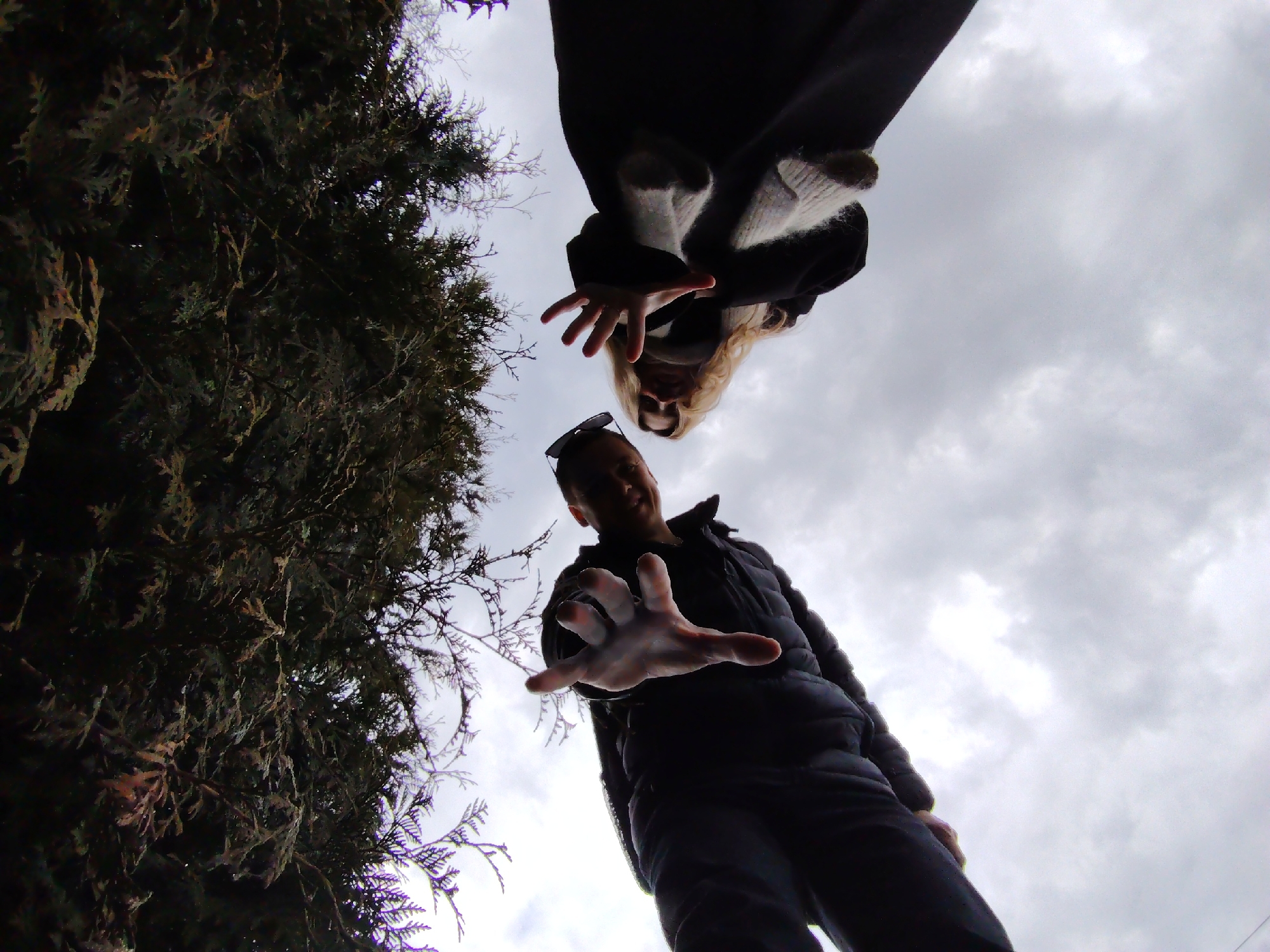 The image shows two people, a man and a woman, from a low angle, with their hands outstretched towards the camera. The man is wearing a dark jacket and sunglasses, while the woman is wearing a dark coat with a light-colored sweater underneath. The sky is overcast with clouds. To the left, there is a tree or shrub. The city of Chaikovsky, Russia is not ascertainable from the available information.